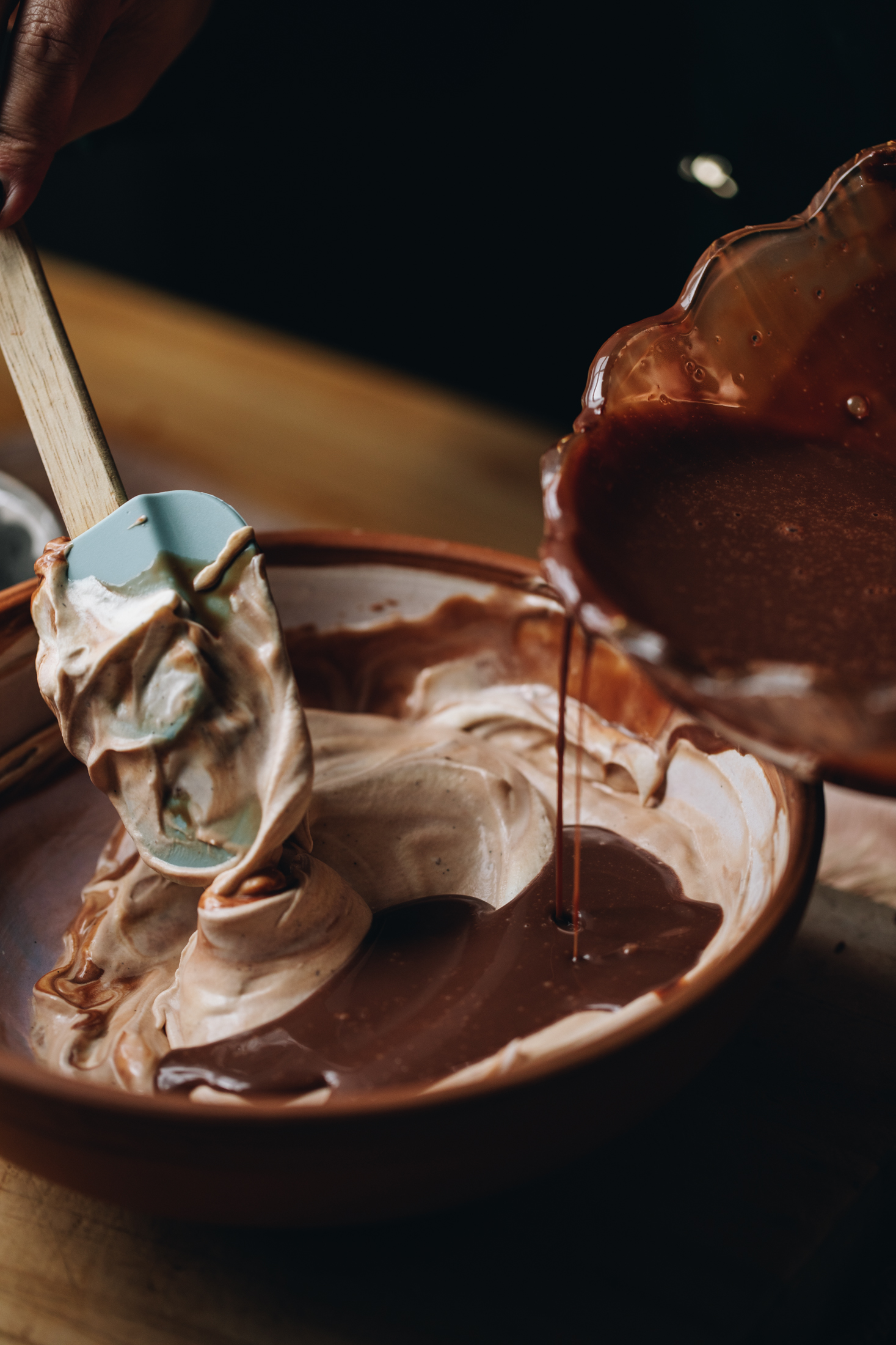 In a ceramic bowl with whipped cream, dark chocolate is being poured in as a blue spatula waits to fold it. It is on a wooden table.