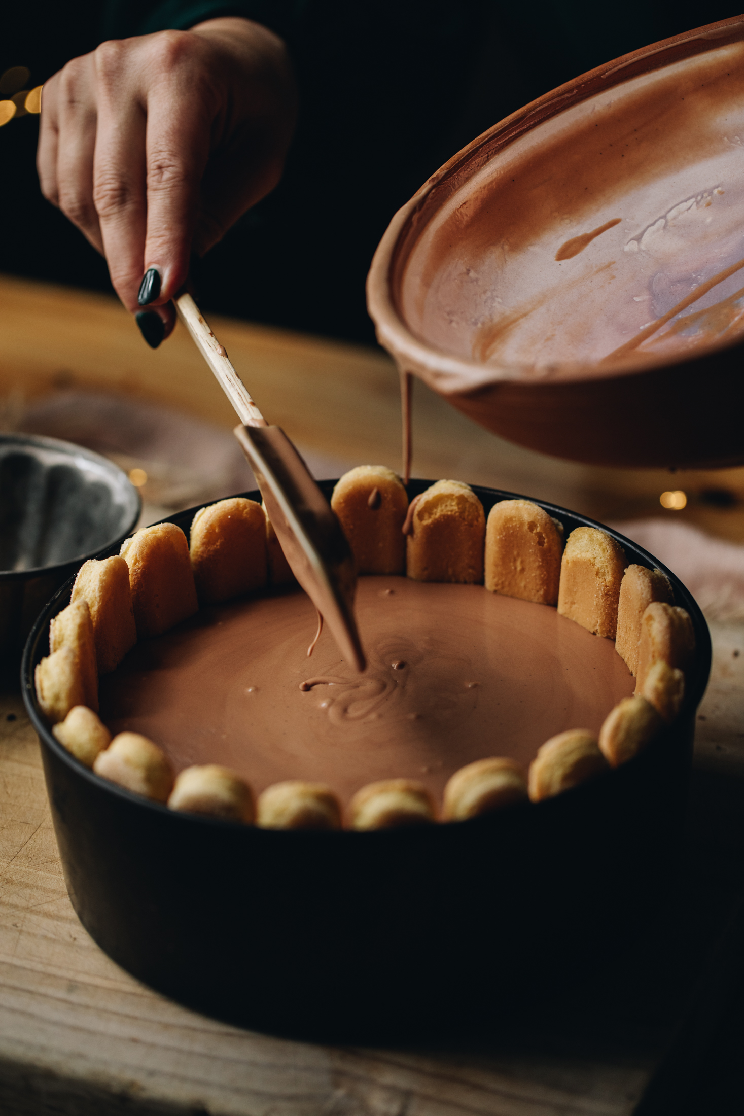A black tin lined with lady fingers has been topped with a milk chocolate cream. It is sitting on a wooden table with spatula ready to smooth the mixture.