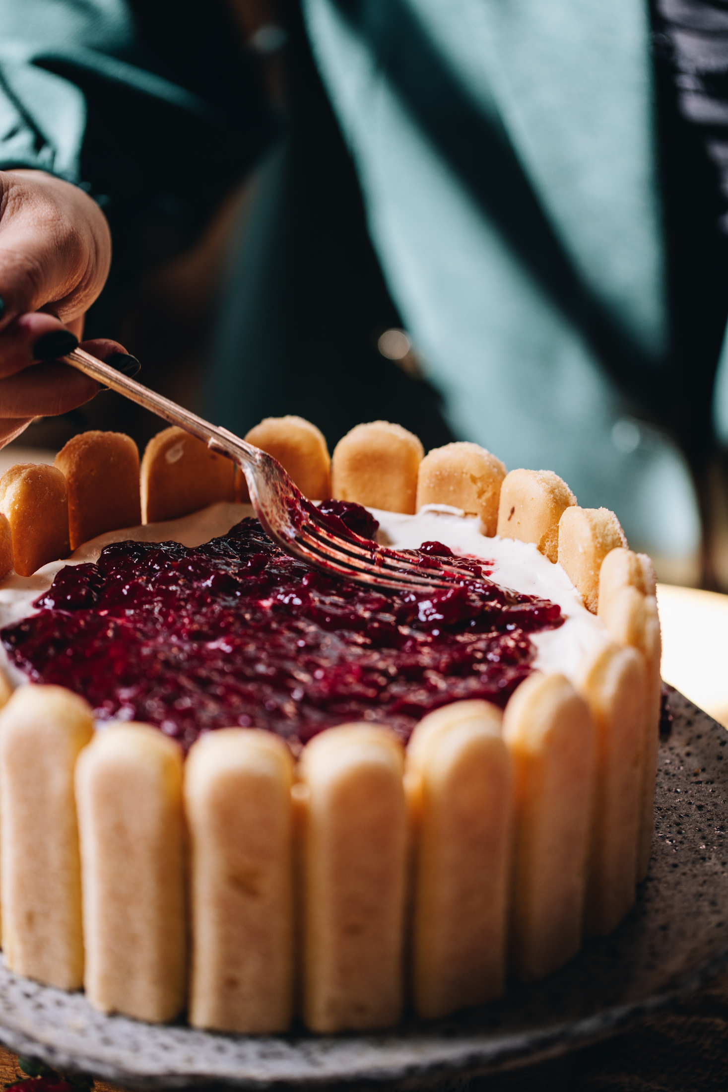 On a ceramic cake stand is a lady finger lined no-bake Chocolate Charlotte Cake. On top is whipped cream and a fork is adding mashed plums that have been smeared on top. 