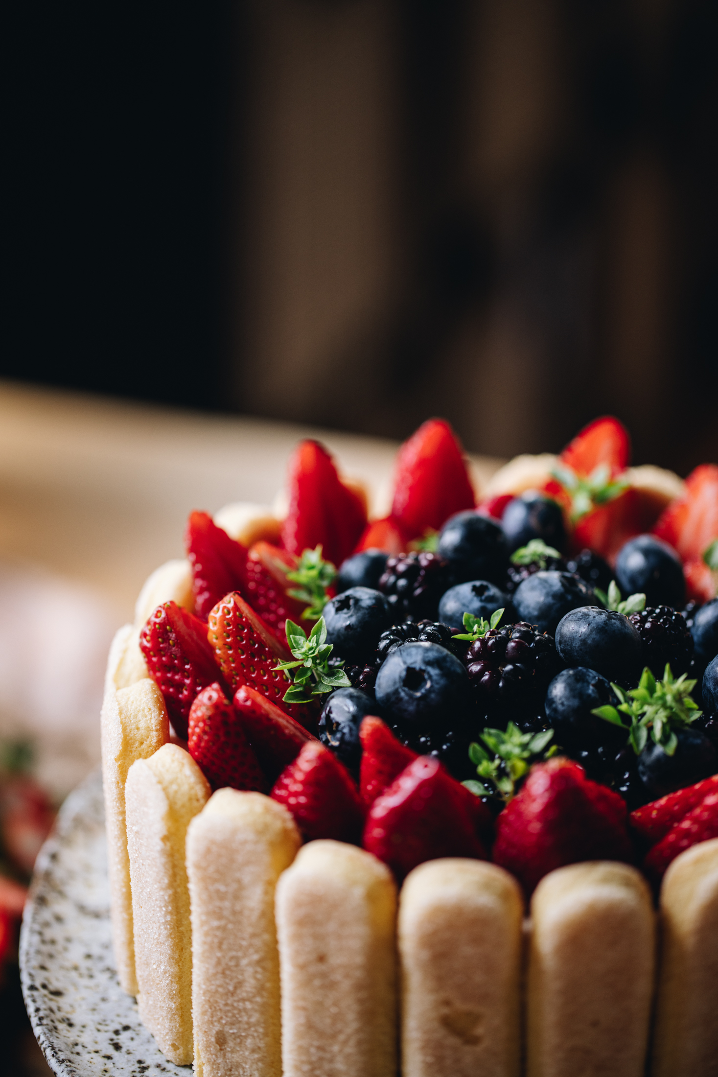 A no-bake Chocolate Charlotte cake is on a ceramic cake stand, on a wooden table. It is decorated with lady fingers around the edge with fresh strawberries on top, blackberries, blueberries and baby basil leaves. 