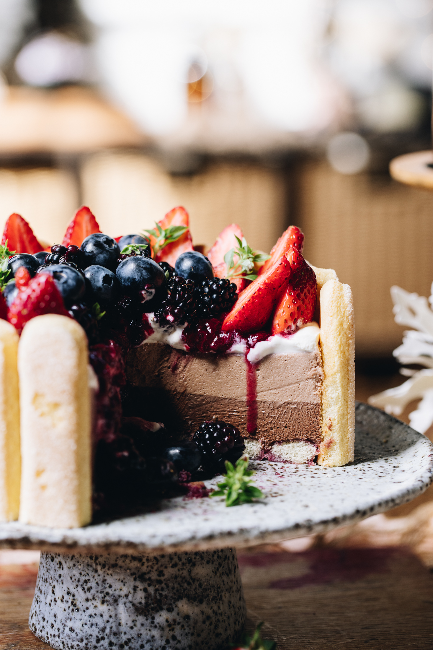 A no-bake Chocolate Charlotte Cake is decorated with berries. It has been cut open to reveal the two tone cake. It is on a ceramic cake stand, on a wooden bench.