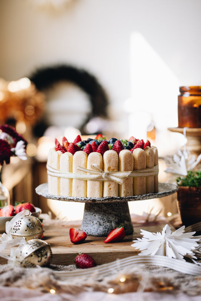 A no-bake Chocolate Charlotte Cake is decorated with berries. It is been tied with a gold ribbon, with a bow at the front. It is on a ceramic cake stand, on a wooden bench. It is surrounded by candles and Christmas decorations. 