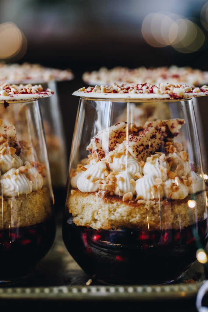 A close up shot shows the Mini Berry Trifles that have been decorated and set in glasses. It shows a pomegranate jelly, a sponge, piped whipped cream, nuts, white chocolate shards. The glass is topped with a white Chocolate disk. 