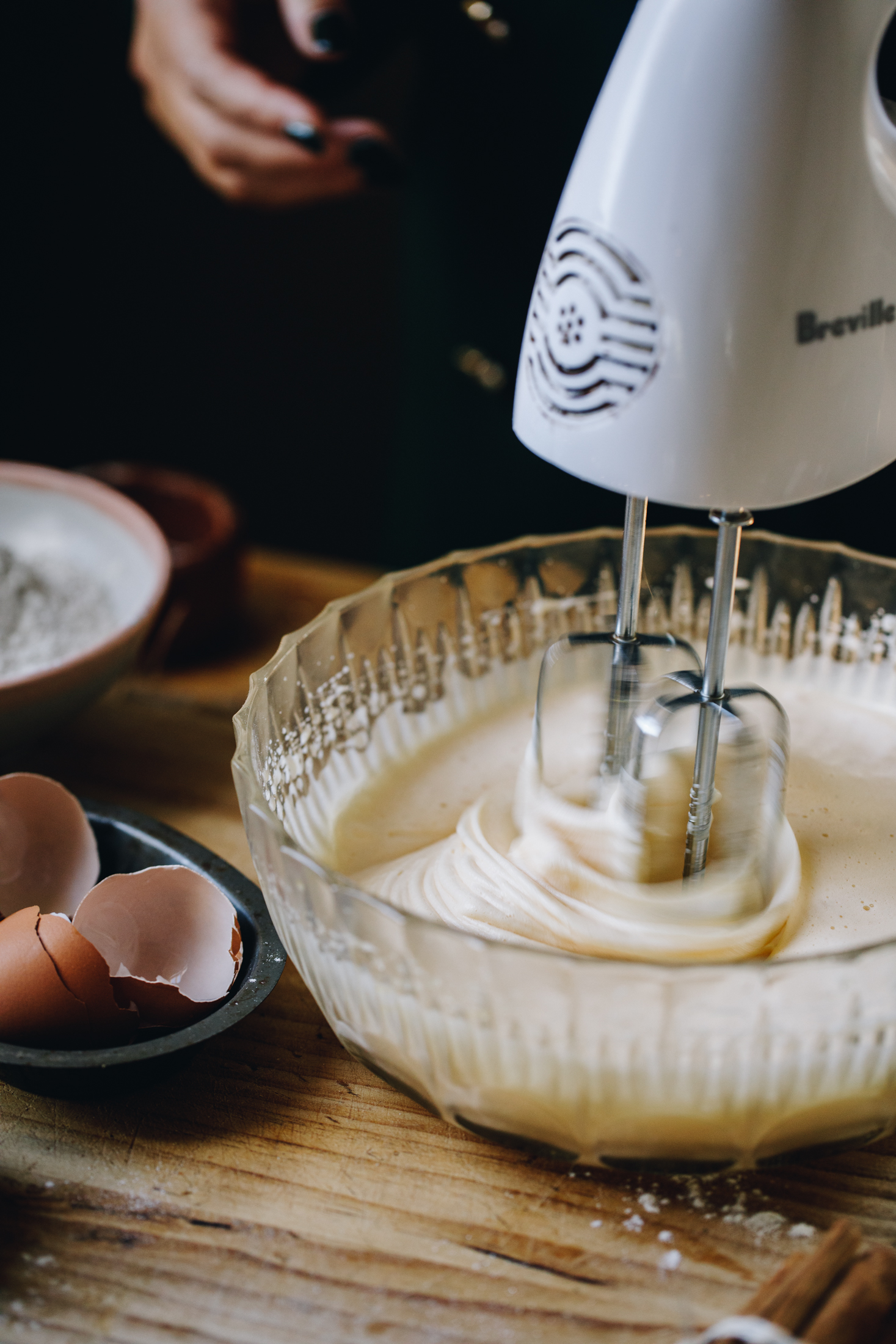 A vintage bowl is on a wooden table. Eggs are being whipped with a white hand mixer. 
