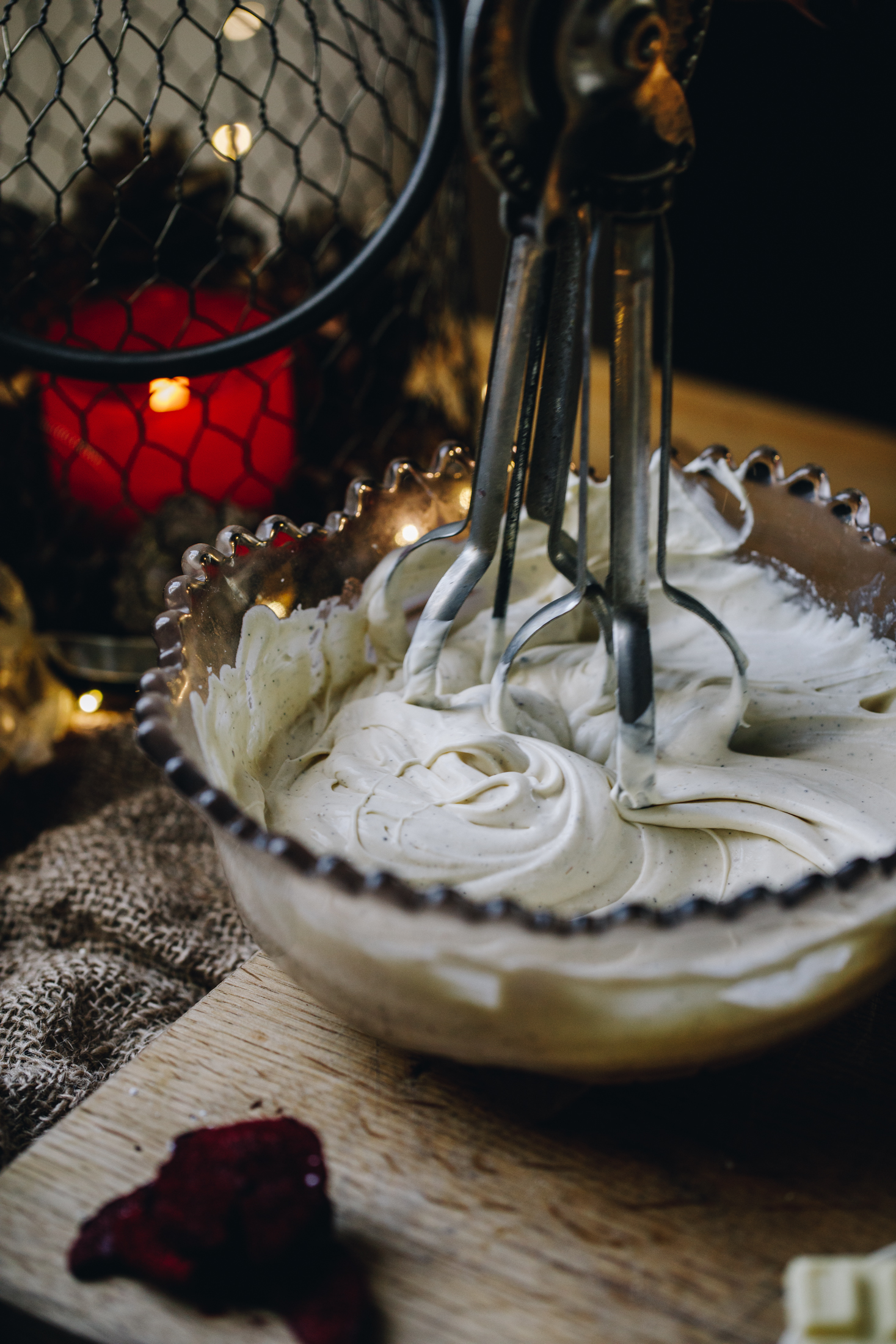 Cream is being whipped in a vintage glass bowl. The cream is thick and ribbony. It is being whipped by a vintage egg beater on a wooden table. An orange candle is in the background. 