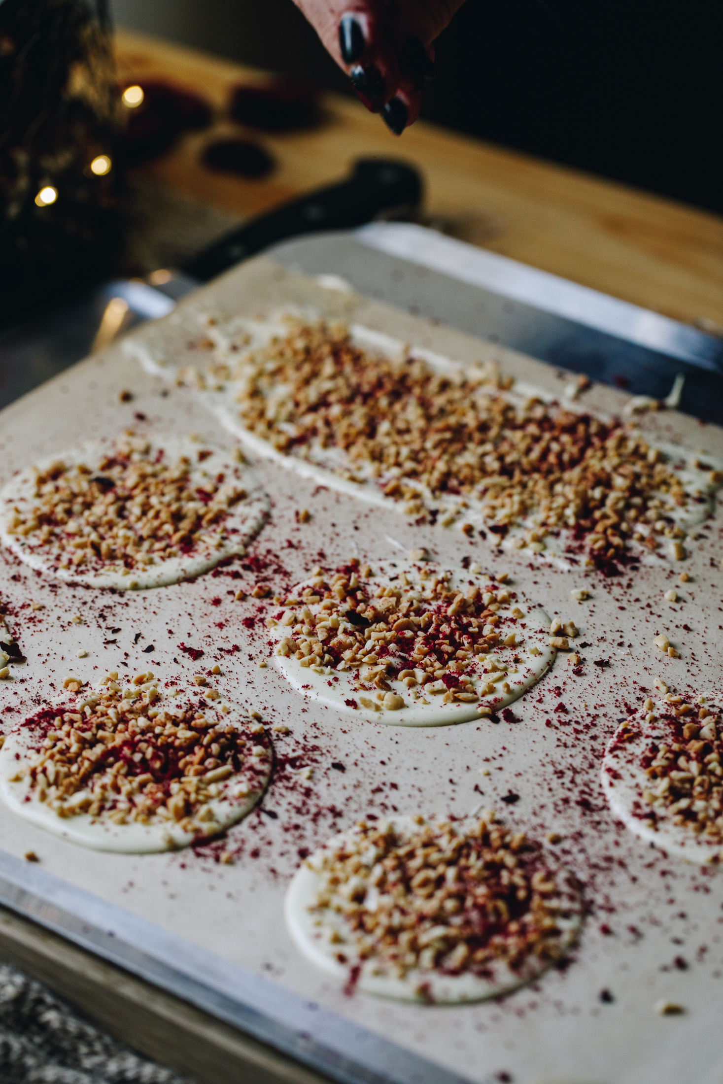 On a silver tray covered with brown paper white chocolate is being smoothed in to discs and a rectangle. All of them are decorated with chopped nuts and freeze-dried boysenberries. 