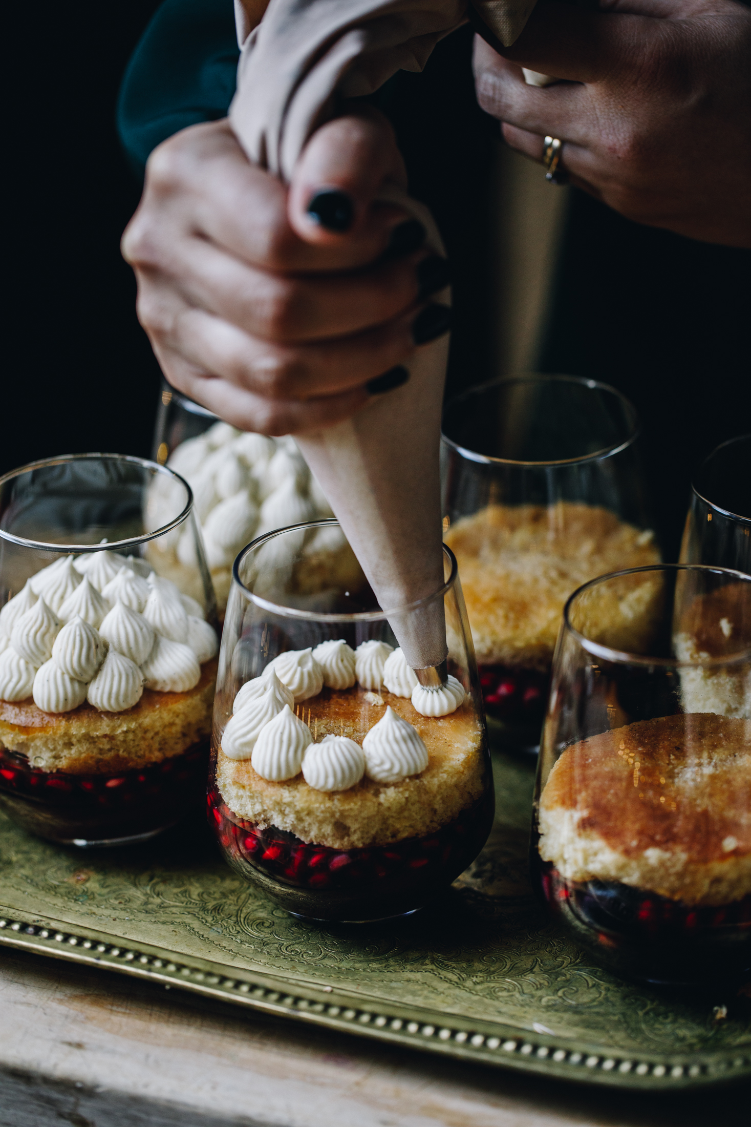 The mini berry trifles have had sponges added to them on top of the set jelly. A sponge is having whipped cream piped on to it. Other glasses already have piles of whipped cream piped on to the sponges. They are on a gold tray. 