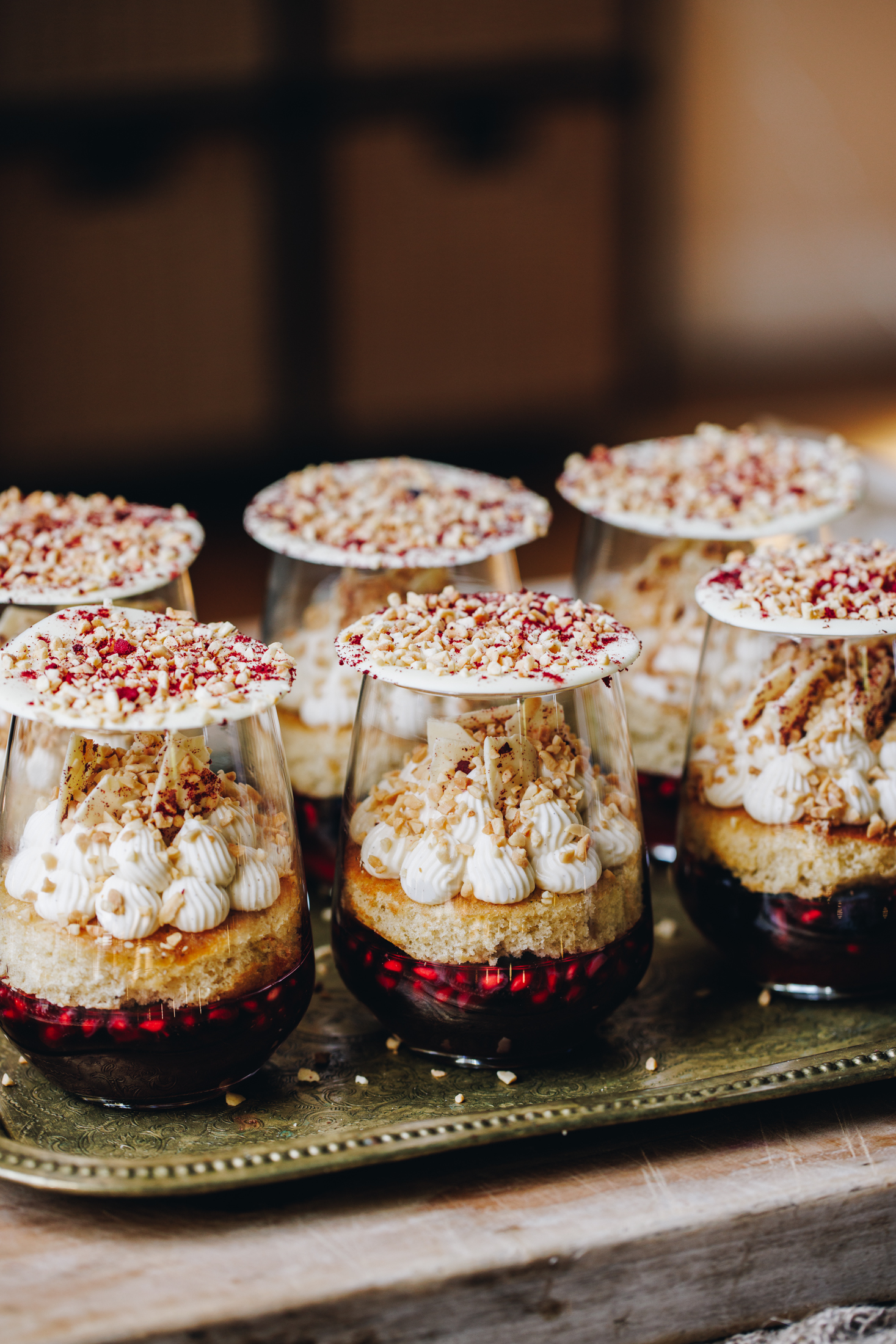 The mini berry trifles have had sponges added to them on top of the set jelly. Sponge has whipped cream piped on to it. The cream has white chocolate shards and chopped nuts on top and white chocolate discs sit over the glasses. 