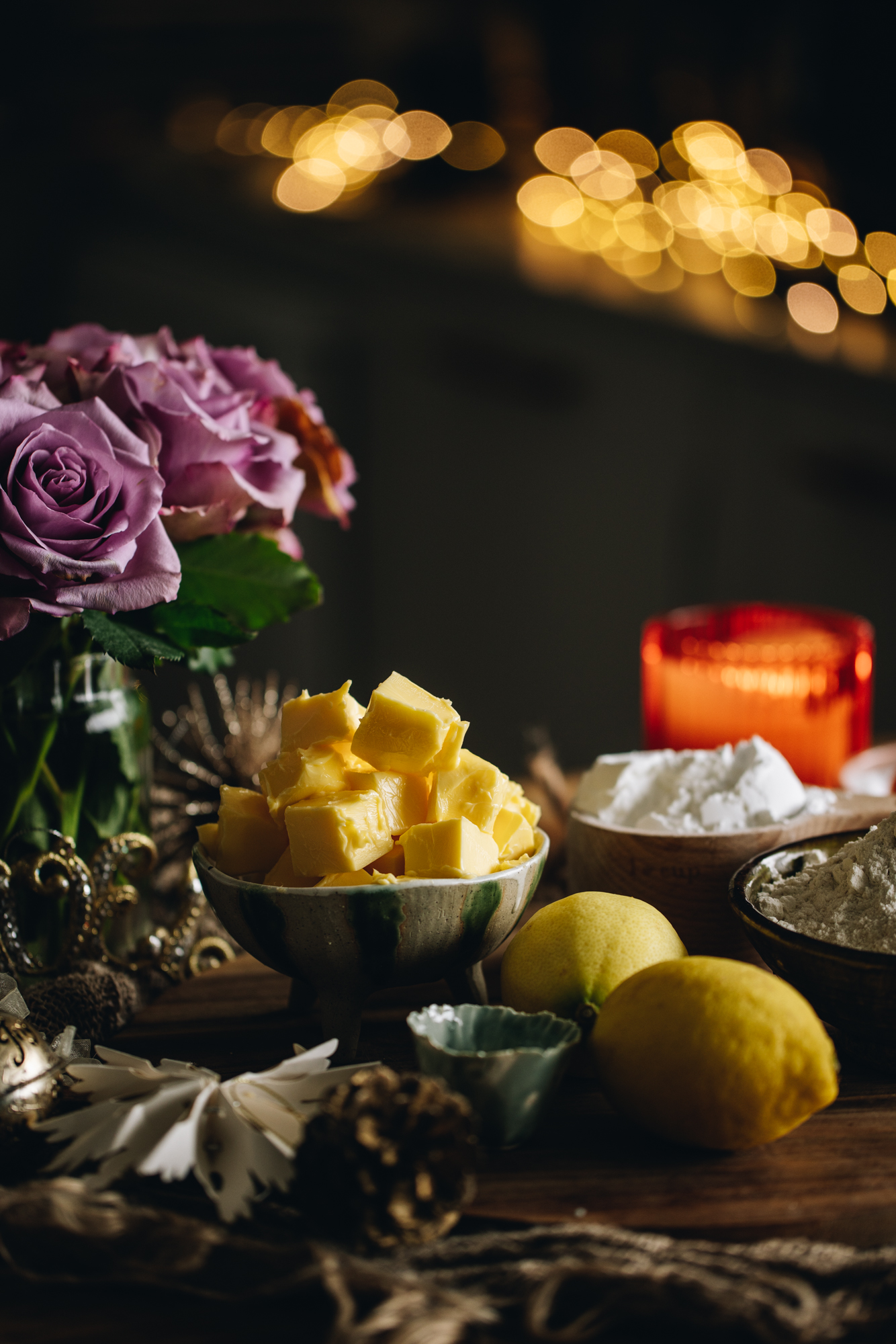 Cookie ingredients are in different vintage bowls and are sitting on a wooden board on top of wooden table There is a bunch of purple flowers next to the ingredients. 