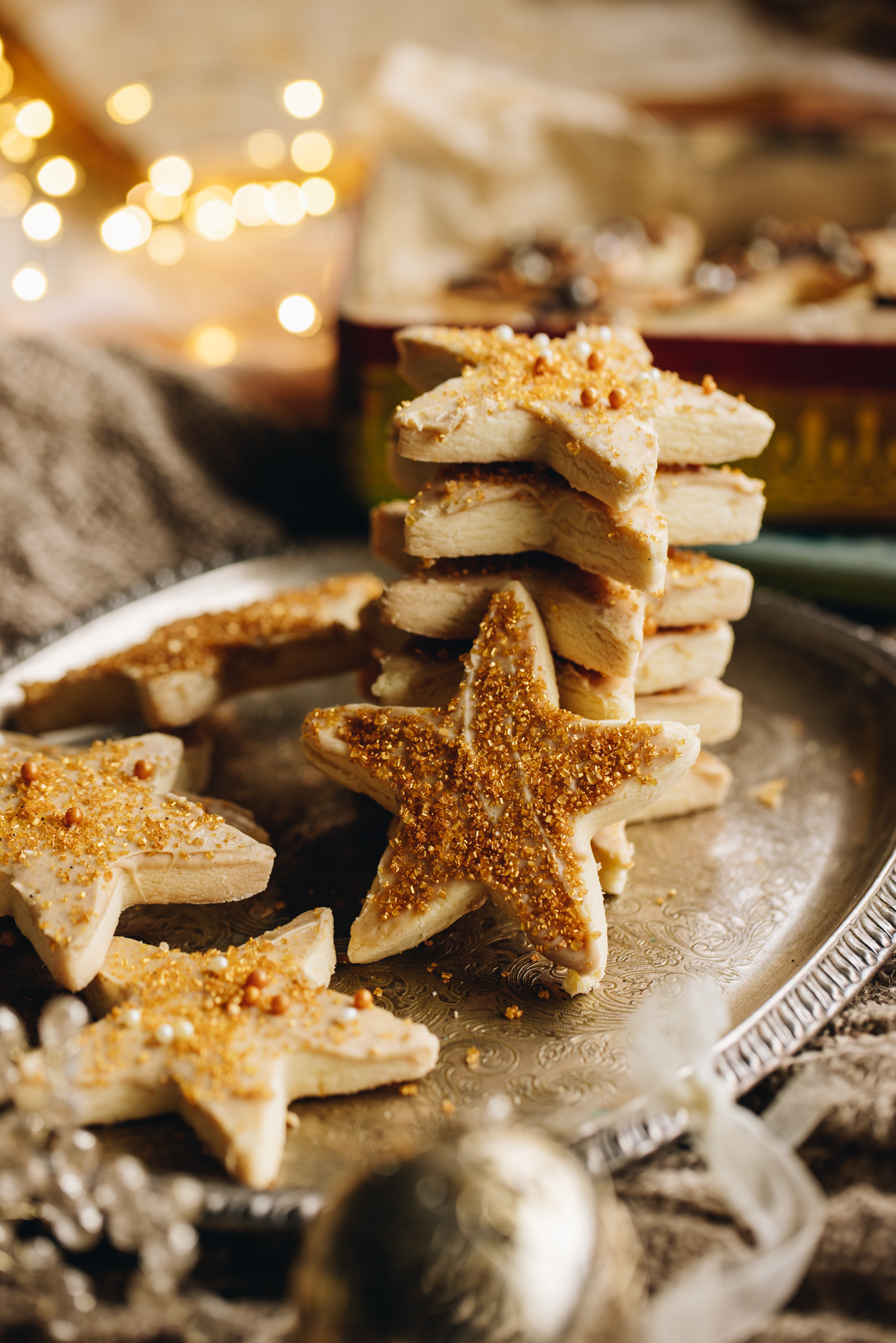 A sliver tray is surrounded by Christmas decorations. On the tray are lemon shortbread cookies that have gold sprinkles and white chocolate underneath them. They are stacked in different angles.