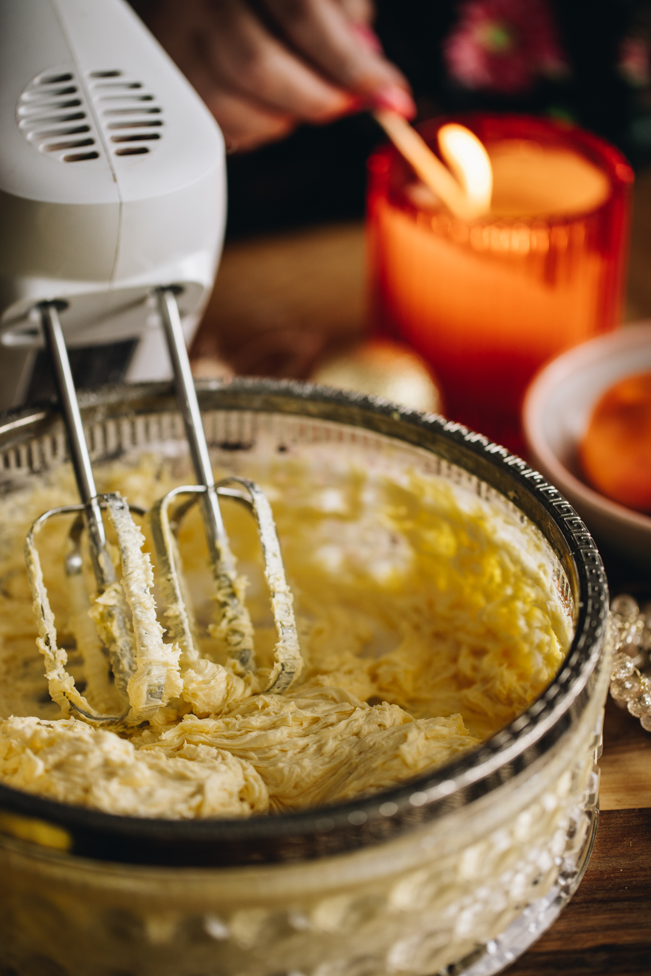 A close of the whipped butter and sugar in a glass bowl that is sitting on a wooden board. An orange candle is being lit in the background. 
