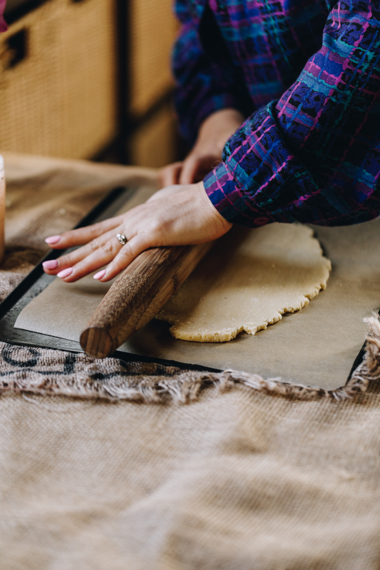On a tablecloth is a silver tray with brown baking paper on it, dough is being rolled out with a wooden rolling pin. 