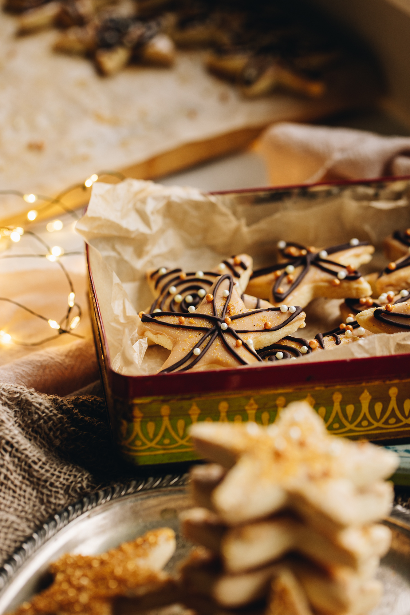In a vintage tin sits freshly decorated lemon shortbread cookies. They are all decorated with white and dark chocolate and orange and white toned sprinkles. 
