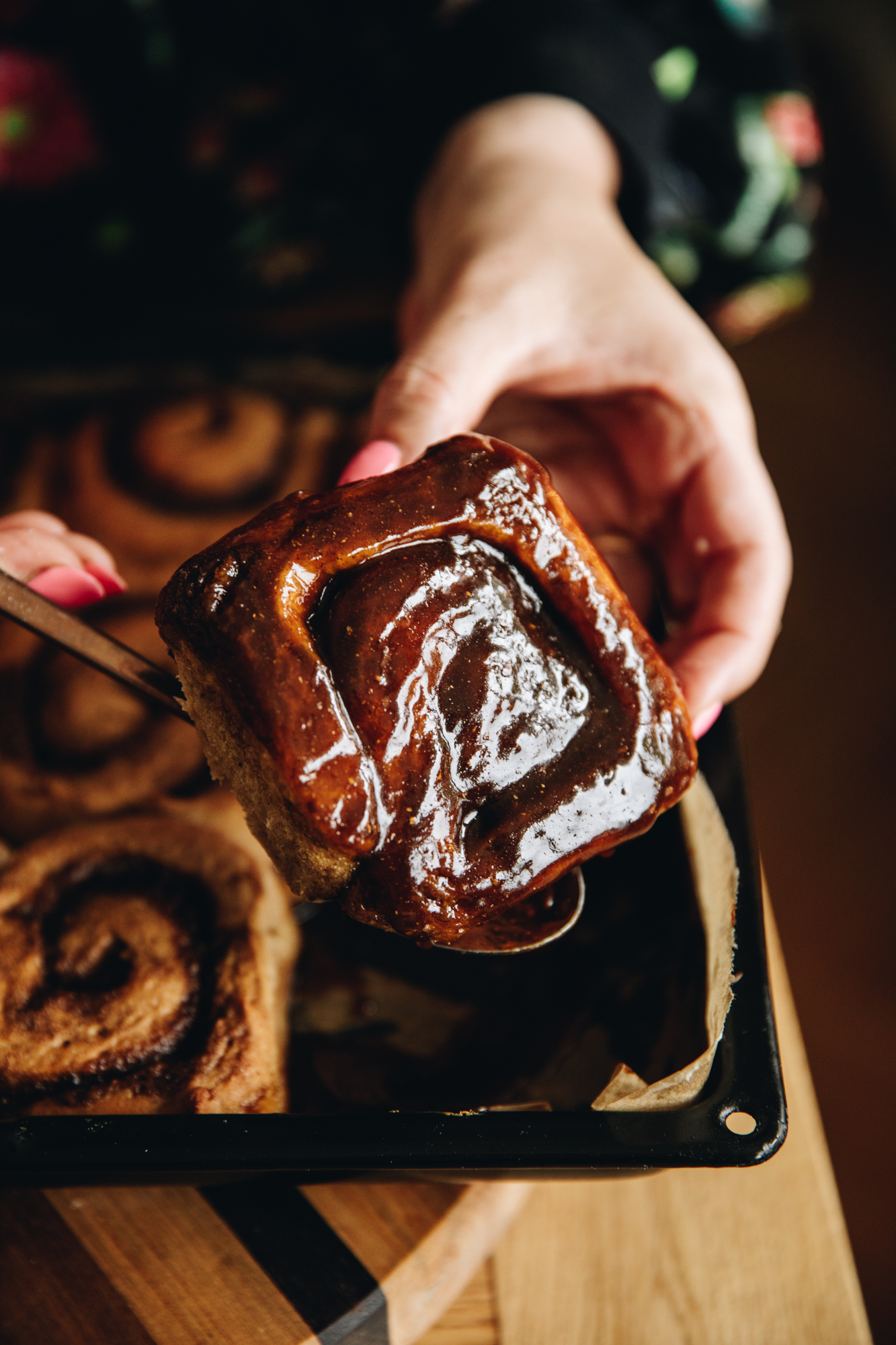 A sticky gingerbread scroll is being lifted out a black tray that is on a wooden board. The scroll is dripping with sticky sauce. 