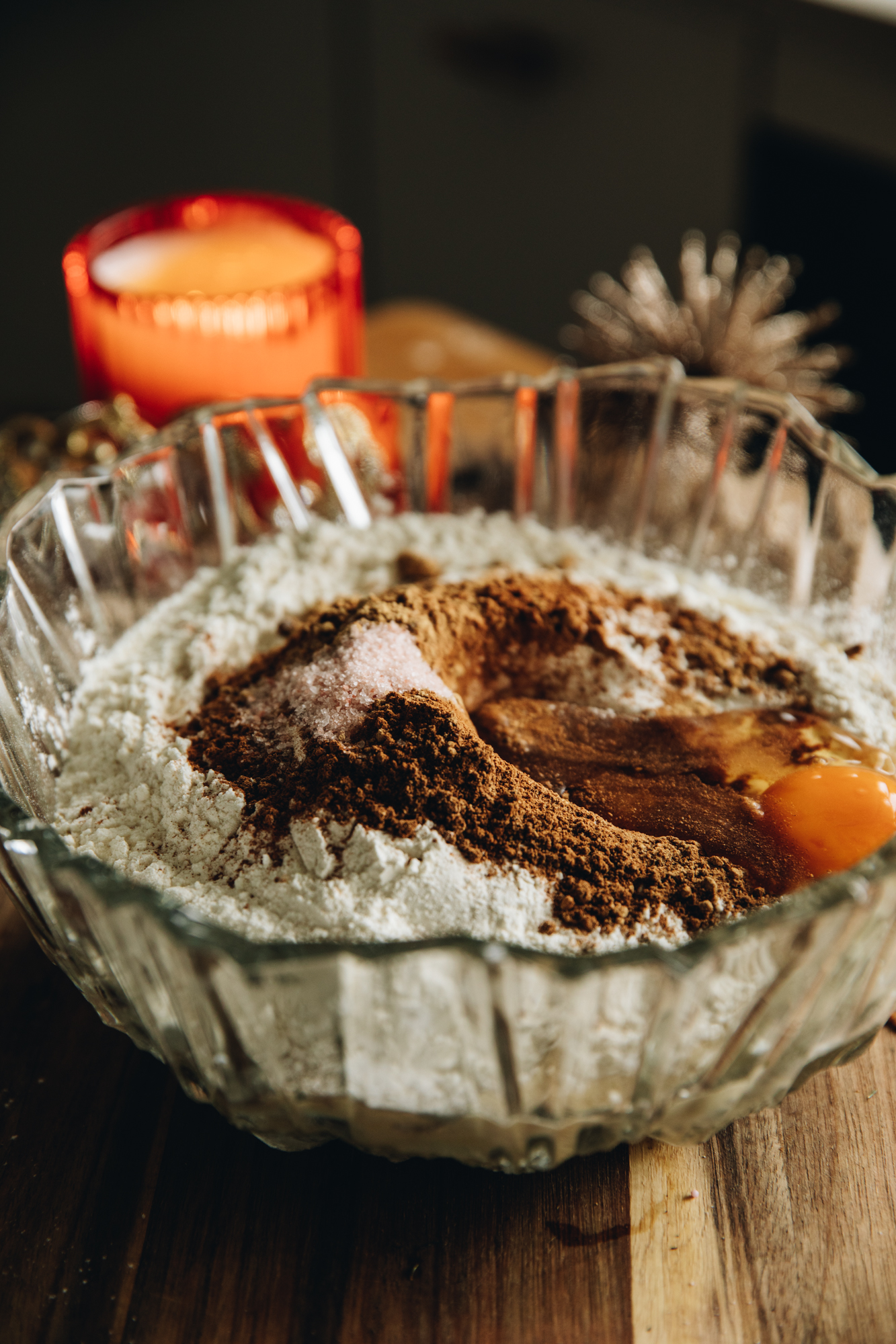 A glass bowl sits a glass bowl on a wooden board. There is an orange candle behind it. In the bowl is flour, spices, salt and an egg.