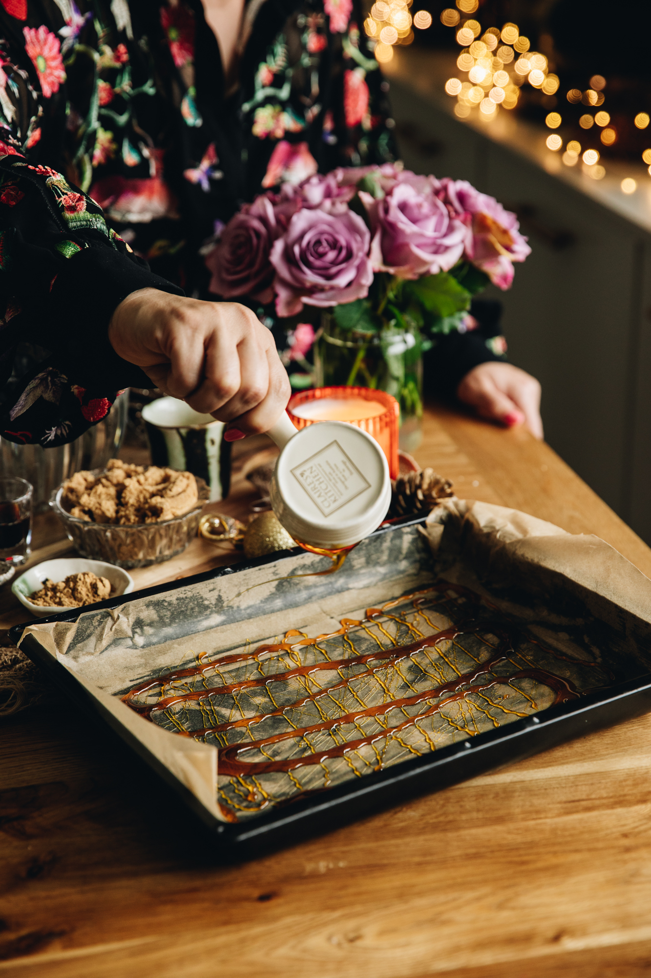 On a wooden table is a black tray lined with baking paper. It is being drizzled with golden syrup. Purple flowers sit in the background and fairy lights can be seen.
