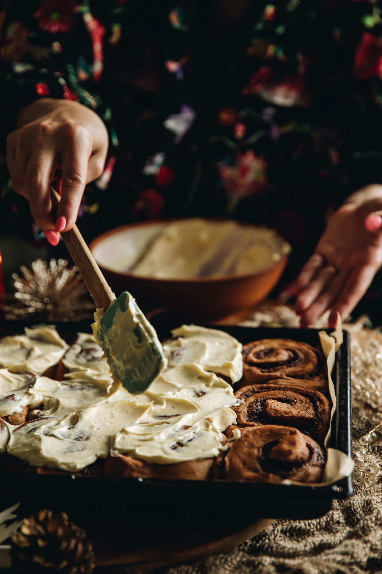 On a black tray are freshly baked sticky gingerbread scrolls. They are being smeared with cream cheese frosting with a spatula.