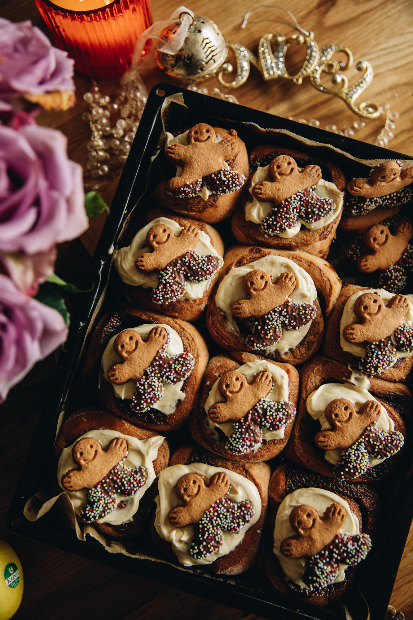 A flat lay shows a black tray are freshly baked sticky gingerbread scrolls. They are topped with cream cheese frosting and a mini gingerbread man are on each scroll.
