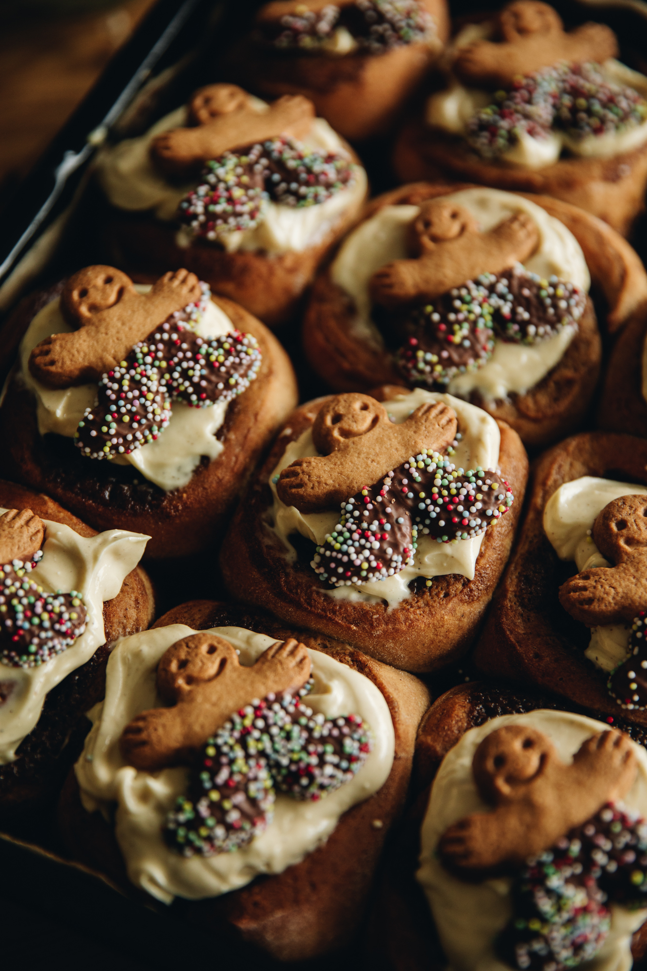 A medium close shot of a black tray of freshly baked sticky gingerbread scrolls. They are topped with cream cheese frosting and are topped with a mini gingerbread man on each scroll.