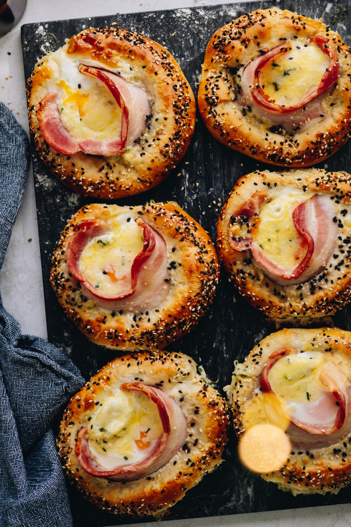 A flat lat shot shows 6 Bacon and Egg Buns that have been freshly baked. The buns have bacon poking out of them with eggs in the centre of each one. The buns are sprinkled with bagel mix, they are on a bdark ceramic board.