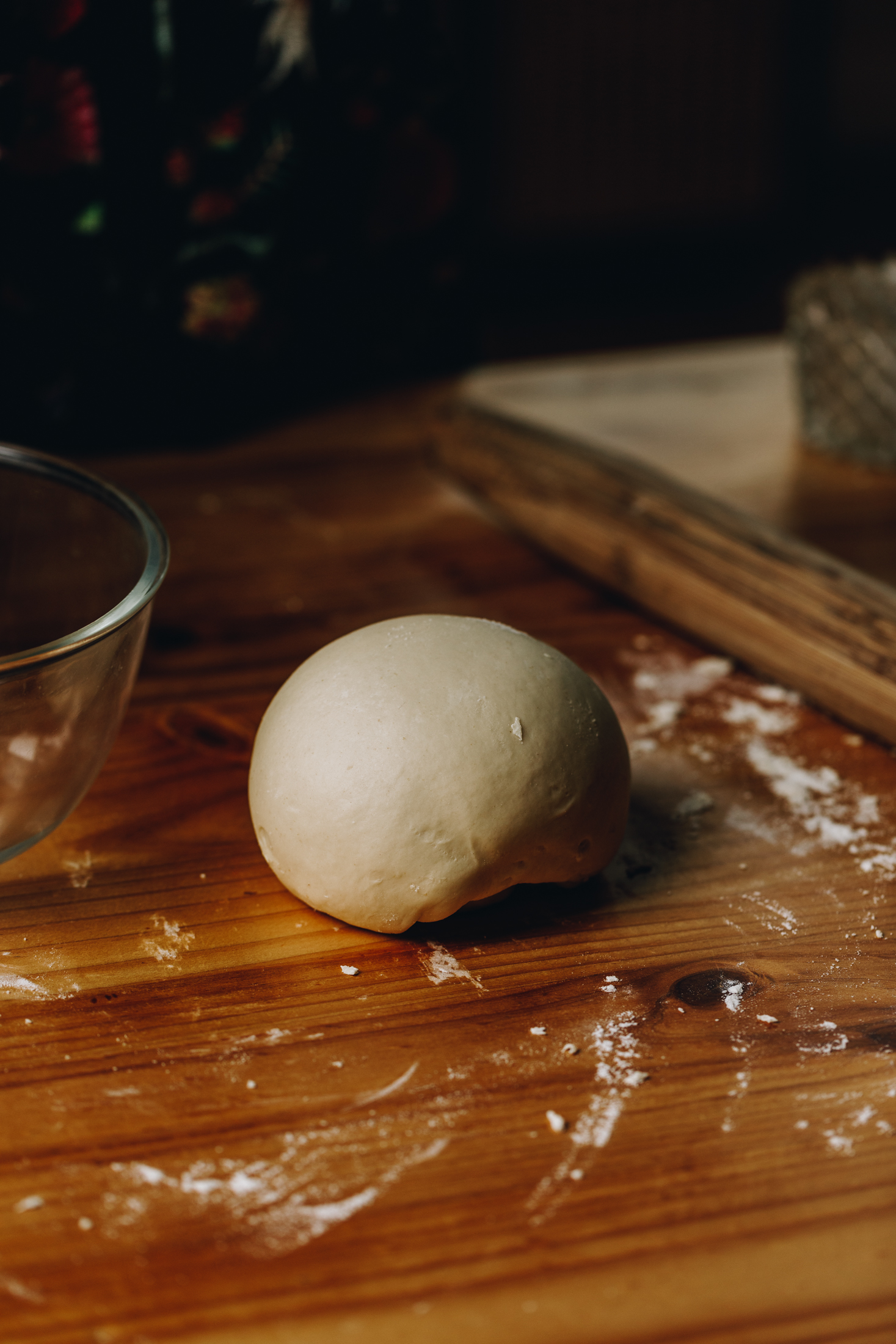 On a floured wooden table is a dough that has been kneaded and is in a ball.