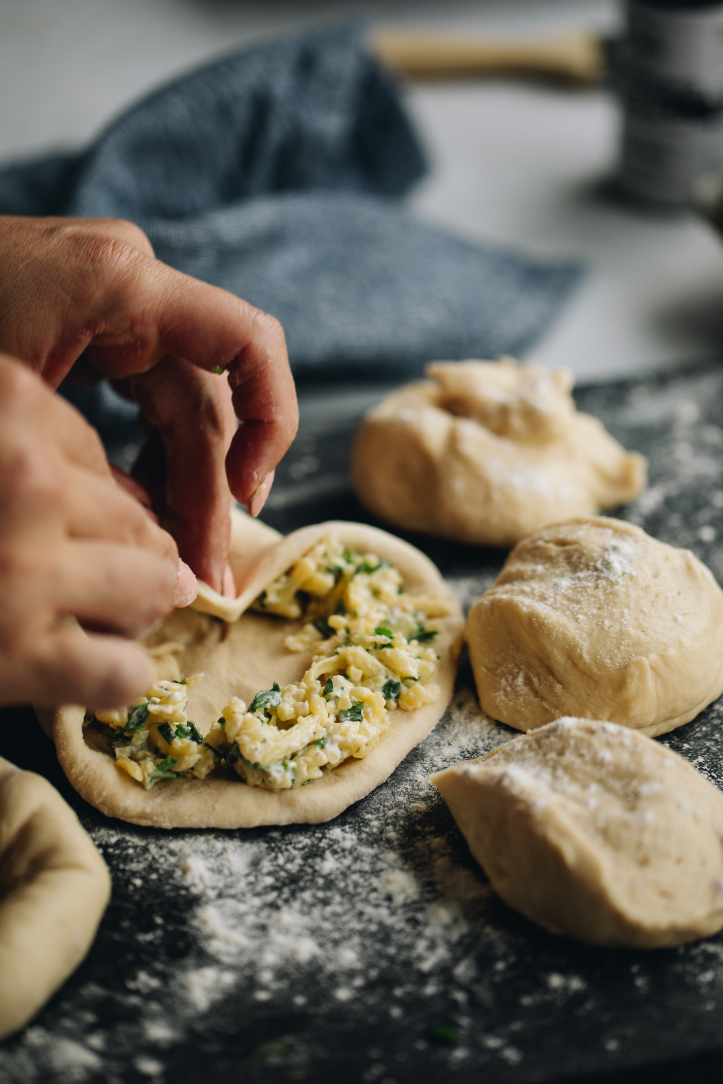 On a blue stone bench are pieces of the Bacon and egg bun dough. A piece of dough has been pressed down and has a herby cheese mixture around the edge and is being encased with the dough.