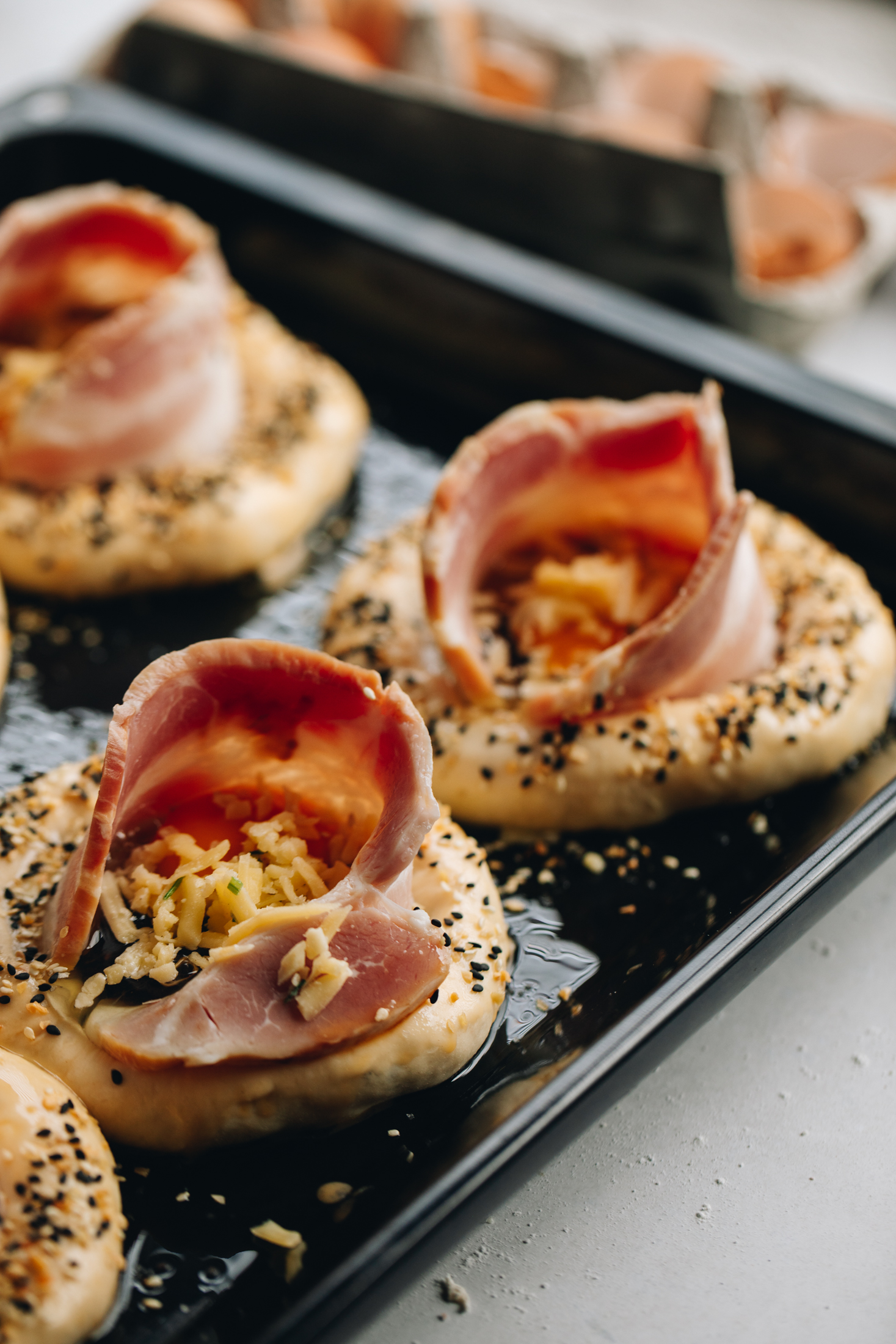 3 Bacon and Egg Buns are unbaked on a black tray. The buns have bacon poking out of them with eggs in the centre of each one. The buns are sprinkled with bagel mix.