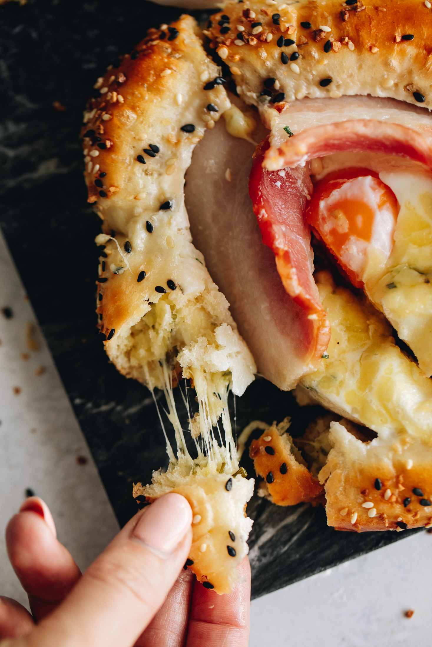 A close up shows one freshly baked bacon and egg bun. A hand is pulling the crust and a cheese pull is seen.