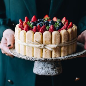 A whole Charlotte Cake is being held in the air by two hands. It is on a ceramic cake stand and is surrounded by lady fingers and topped with fresh berries.