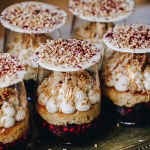 A tray of mini berry trifles are on a gold tray. They are filled with red jelly, sponge, piped cream, chocolate shards and white chocolate discs sit on top of each glass.