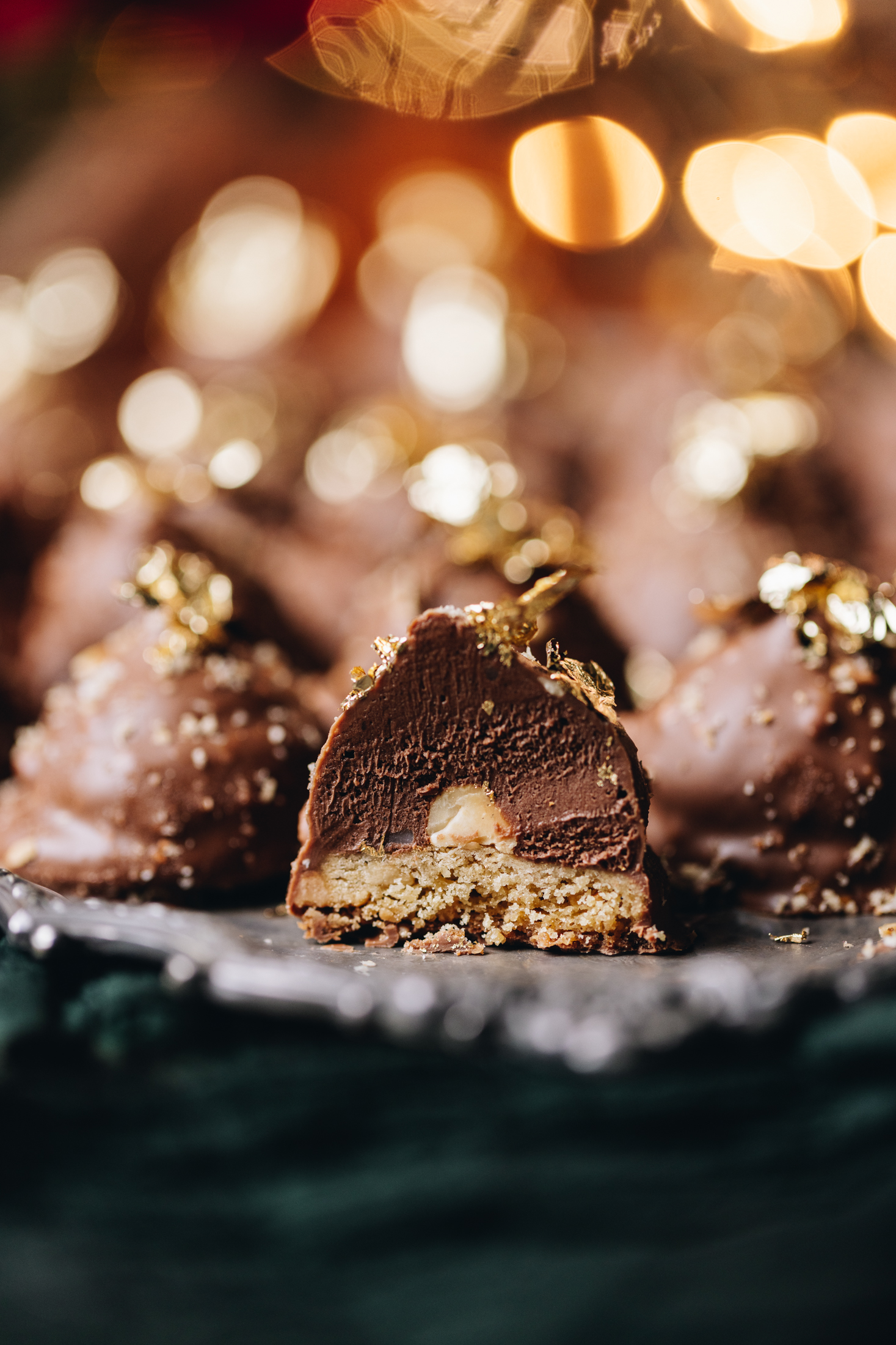 A silver tray sits on top of a green cloth. On top of the tray are freshly baked chocolate hazelnuts cookies. One of them at the front has been cut open to reveal the hazelnut cookie base, the creamy Nutella filling with a hazelnut inside, topped with chocolate, hazelnut crumb and gold leaf. There are fairy lights in the background.