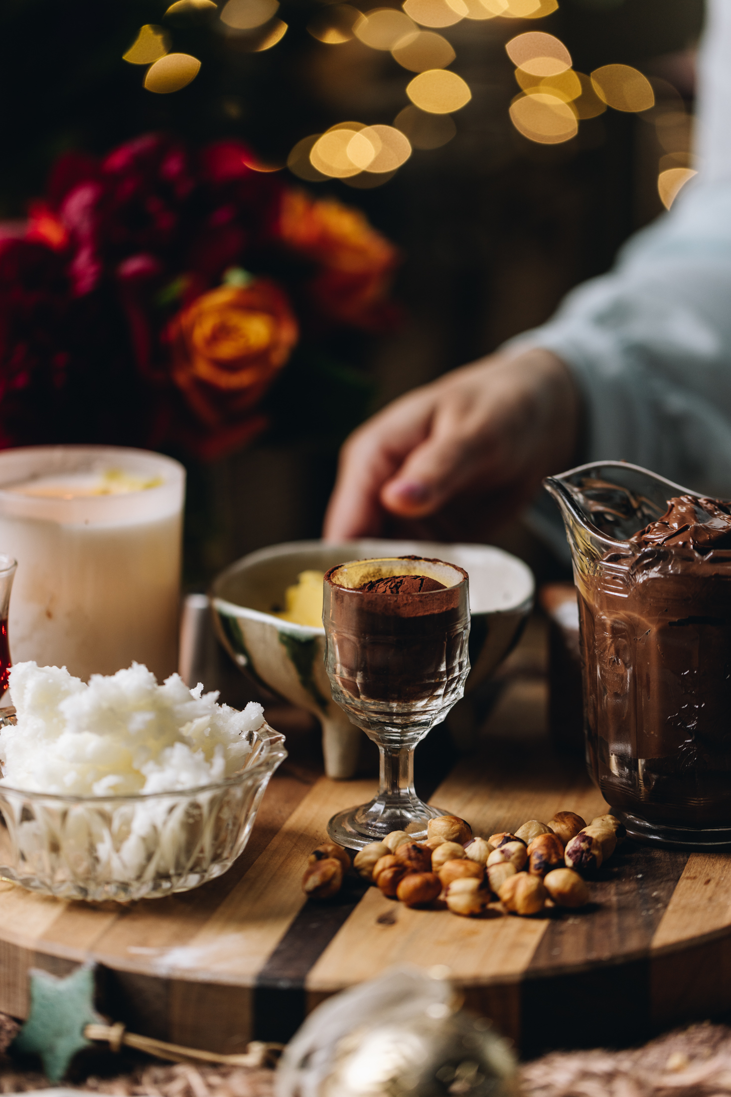 A wooden board is filled with biscuit ingredients. There is coconut oil, cocoa, Nutella, toasted hazelnuts and butter in shot. A burning candle and flowers are in the background with fairy lights blurred in the distance. Naomi's hand is reaching to the butter to grab it.