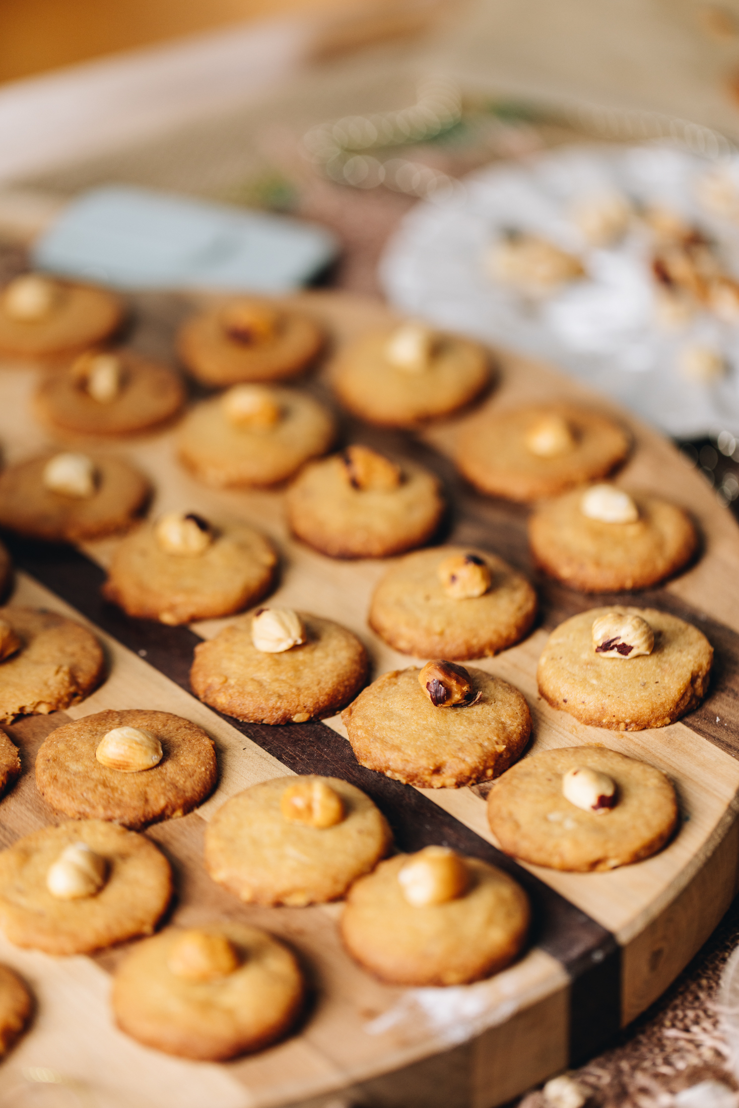 A striped wooden board sits on top of natural fabric. On the board is freshly baked hazelnut cookies that have a hazelnut placed in the centre of each one. A blue spatula is in the background.