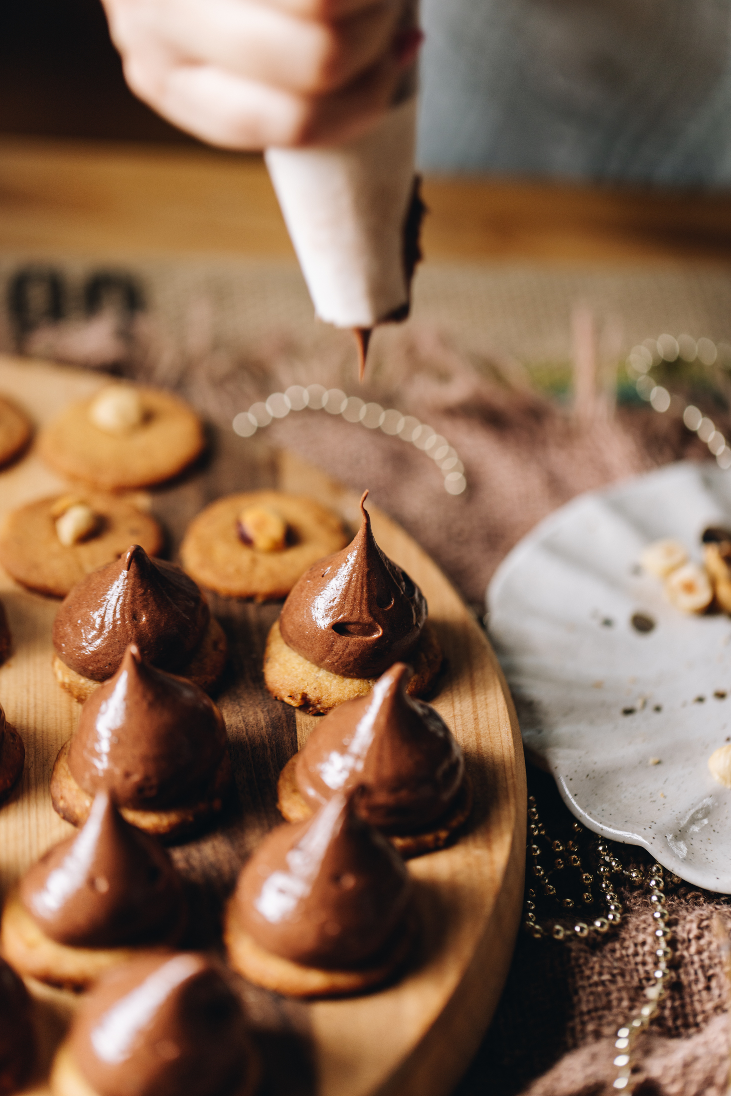 A striped wooden board sits on top of natural fabric. On the board is freshly baked hazelnut cookies that have a hazelnut placed in the centre of each one. Nutella chocolate filling has been piped on to seven on the cookies and one has just been piped with a pointy tip. A ceramic plate with a few hazelnuts is in the background.