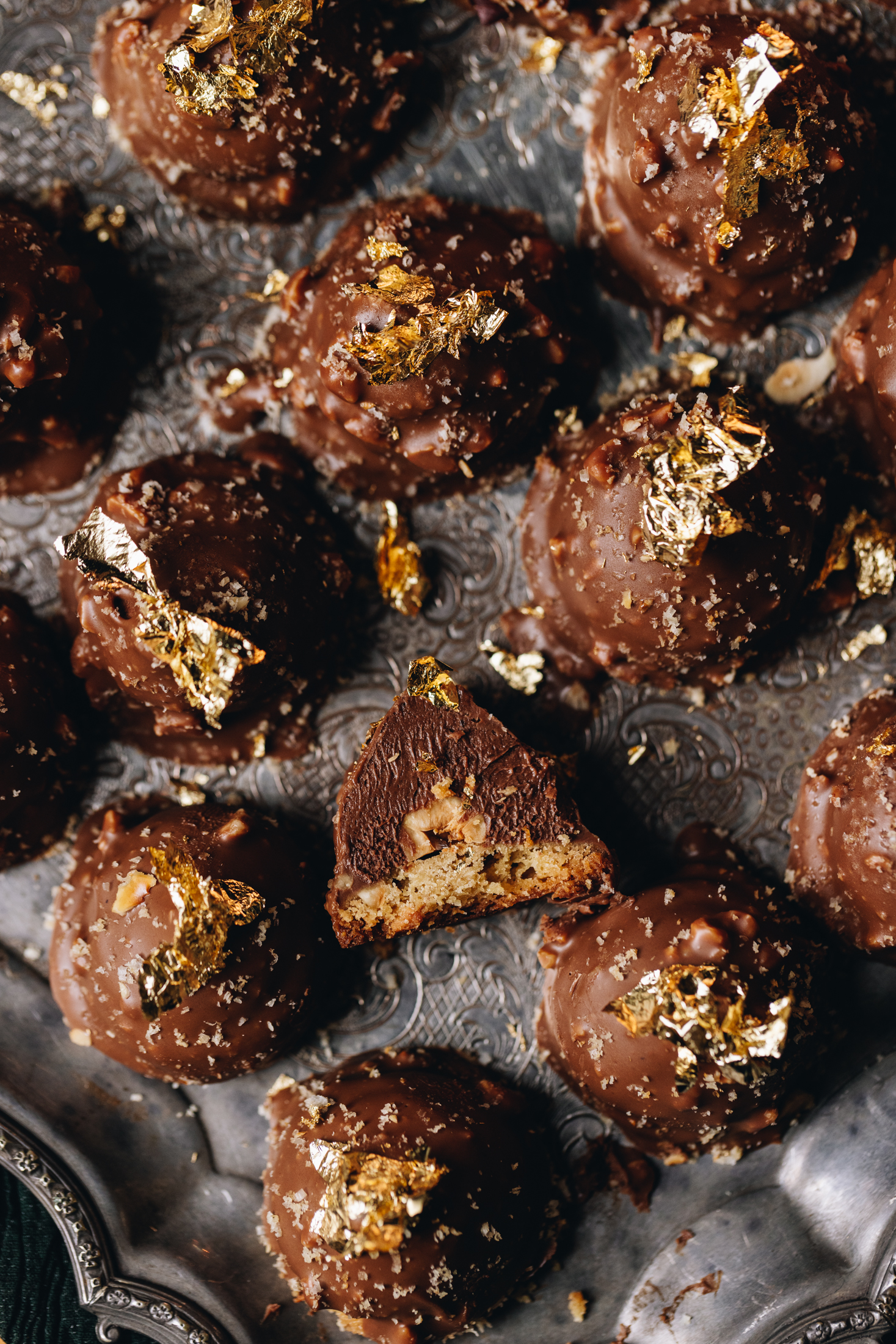 On top of a vintage silver tray are freshly baked chocolate hazelnuts cookies. One of them at the front has been cut open to reveal the hazelnut cookie base, the creamy Nutella filling with a hazelnut inside, topped with chocolate, hazelnut crumb and gold leaf. It is a flatly shot.
