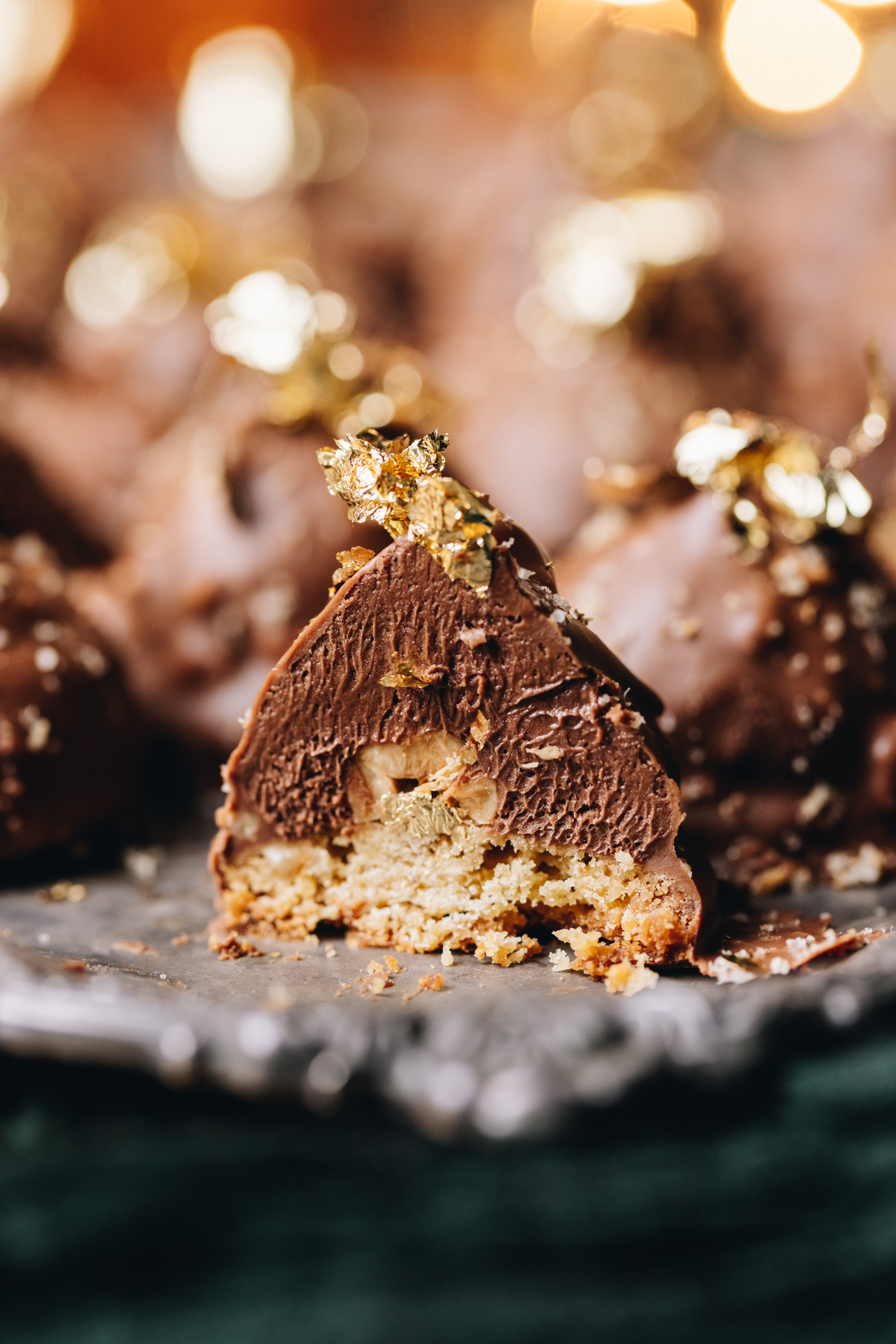 A silver tray sits on top of a green cloth. On top of the tray are freshly baked chocolate hazelnuts cookies. One of them at the front has been cut open to reveal the hazelnut cookie base, the creamy Nutella filling with a hazelnut inside, topped with chocolate, hazelnut crumb and gold leaf. There are fairy lights in the background.