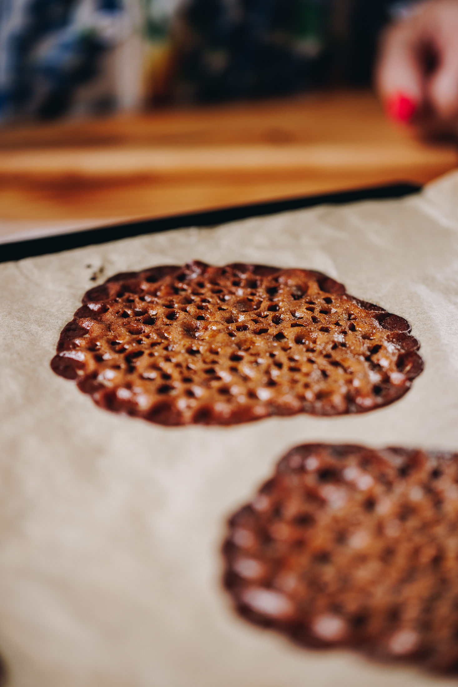Two freshly baked brandy snaps are on brown paper, on a wooden table. They are flat and unrolled. 
