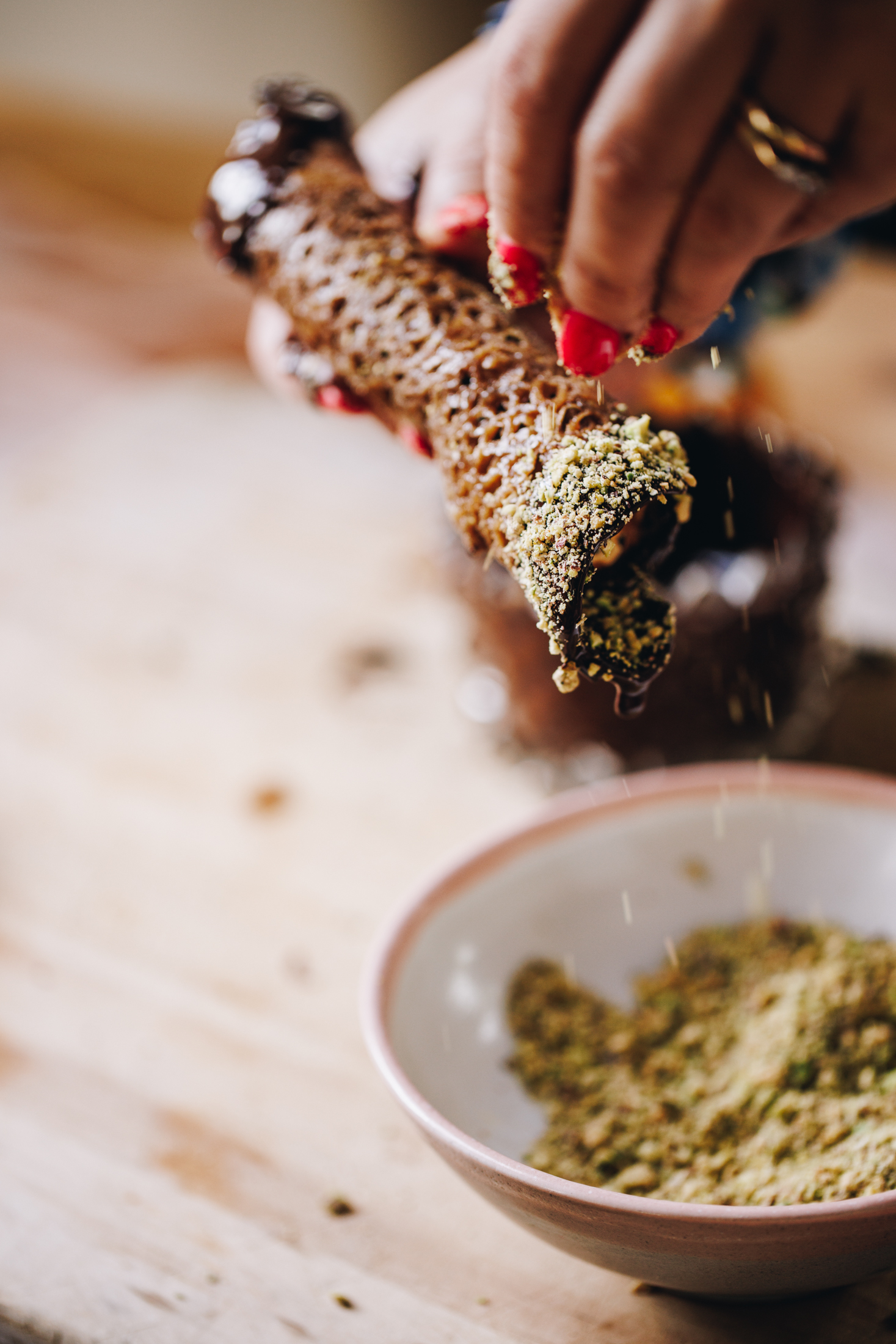 A Brandy snap is being sprinkled with pistachios crumbs, on top of a chocolate dipped edge. The bowl of pistachios is on a wooden table. 