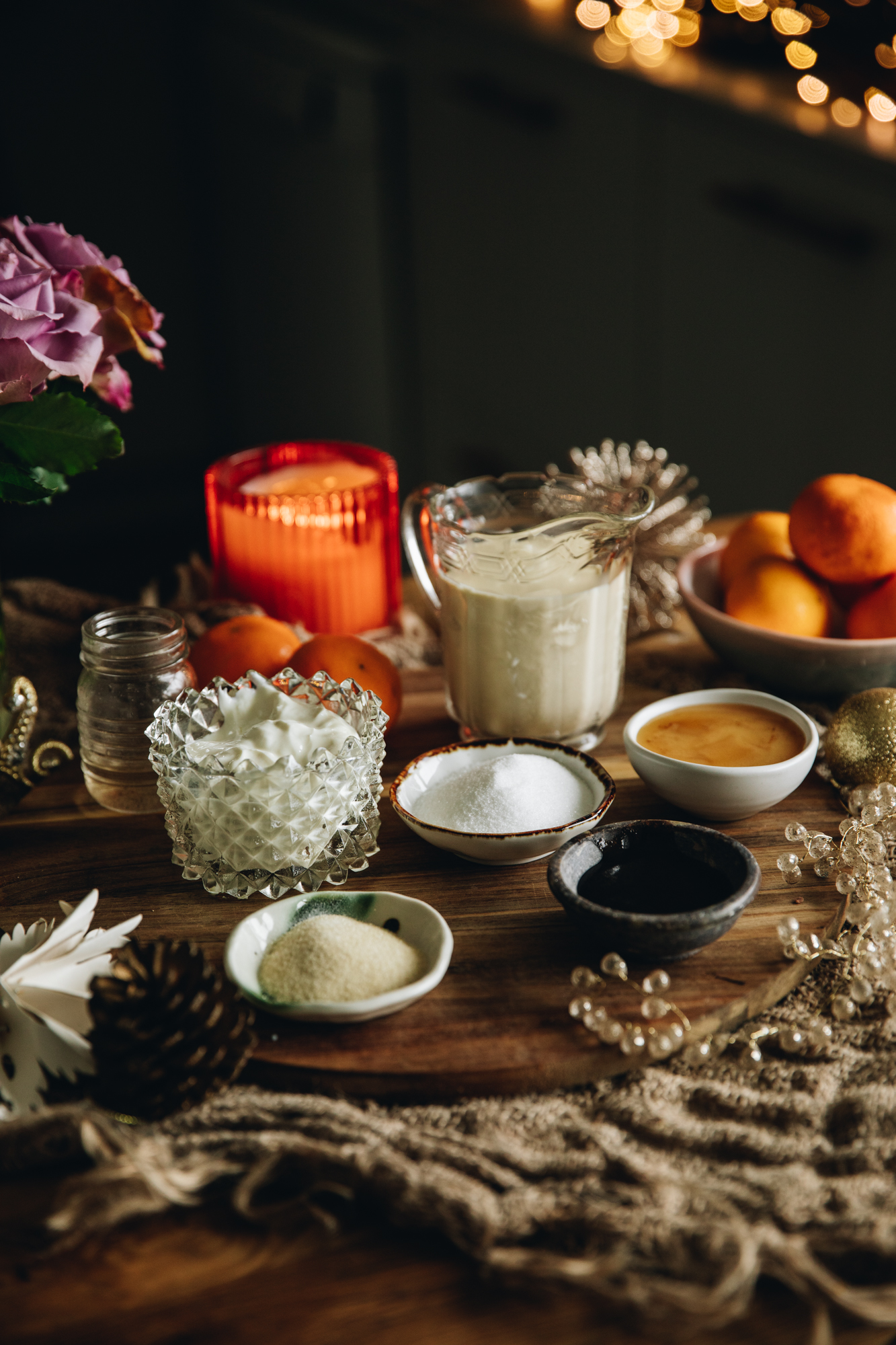 On a wooden board sits vintage bowls and small plates that have pan cotta ingredients on it. There is cream, honey, oranges and sugar in view. They are sitting on a wooden table with natural fabrics on it, an orange candle and purple flowers are in the background.
