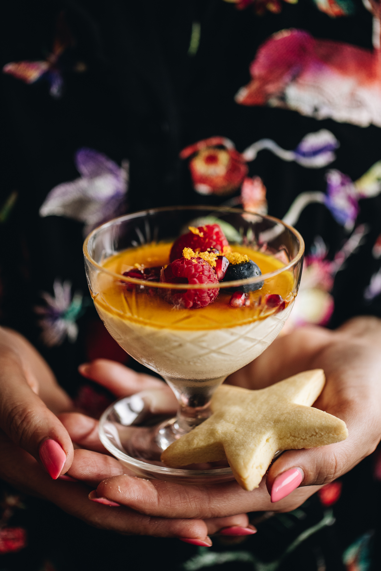 A single Orange Panna Cotta is being held in Naomi's hands. It is a vintage glass with the creamy panna cotta and orange syrup on top of it with fresh raspberries and blueberries. There is a plain star shortbread biscuit next to it. 
