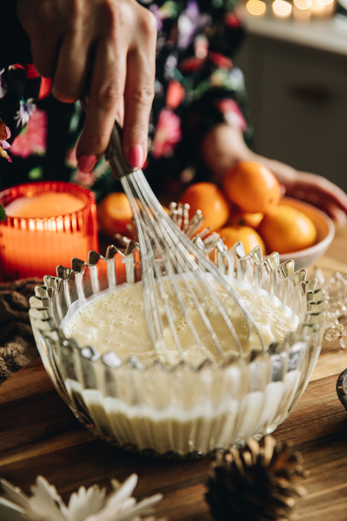 A glass vintage bowl sits on a wooden table filled with cream. An orange candle burns in the background along with a bowl of mandarins and natural fabrics.