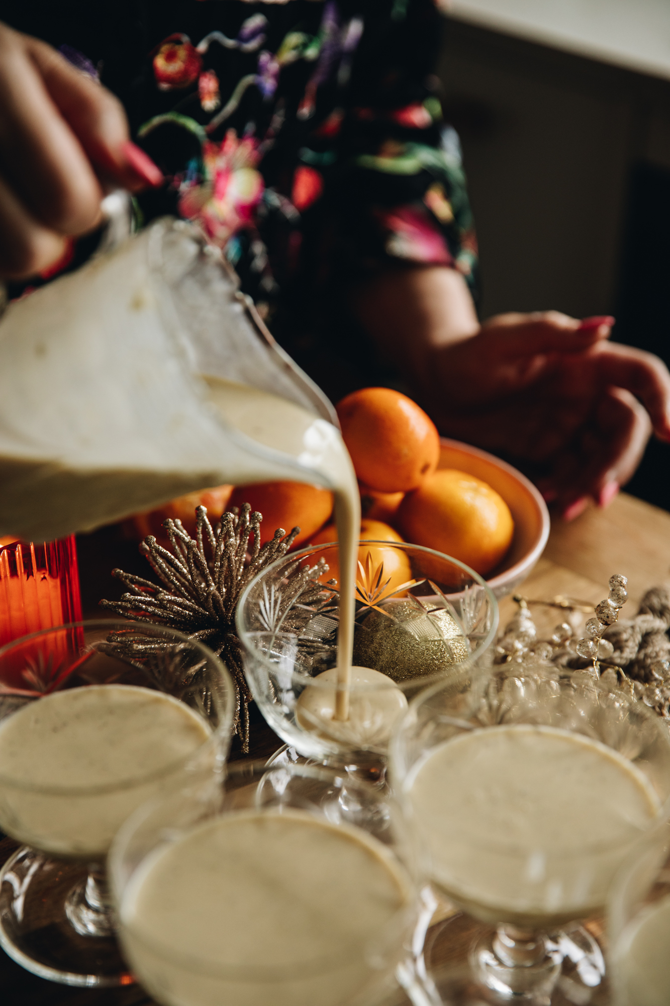 4 glass vintage ramekins sit on a wooden table. Three of them have been filled with the cream mixture and vintage glass jug is adding the cream to the fourth.. An orange candle burns in the background along with a bowl of mandarins and natural fabrics.