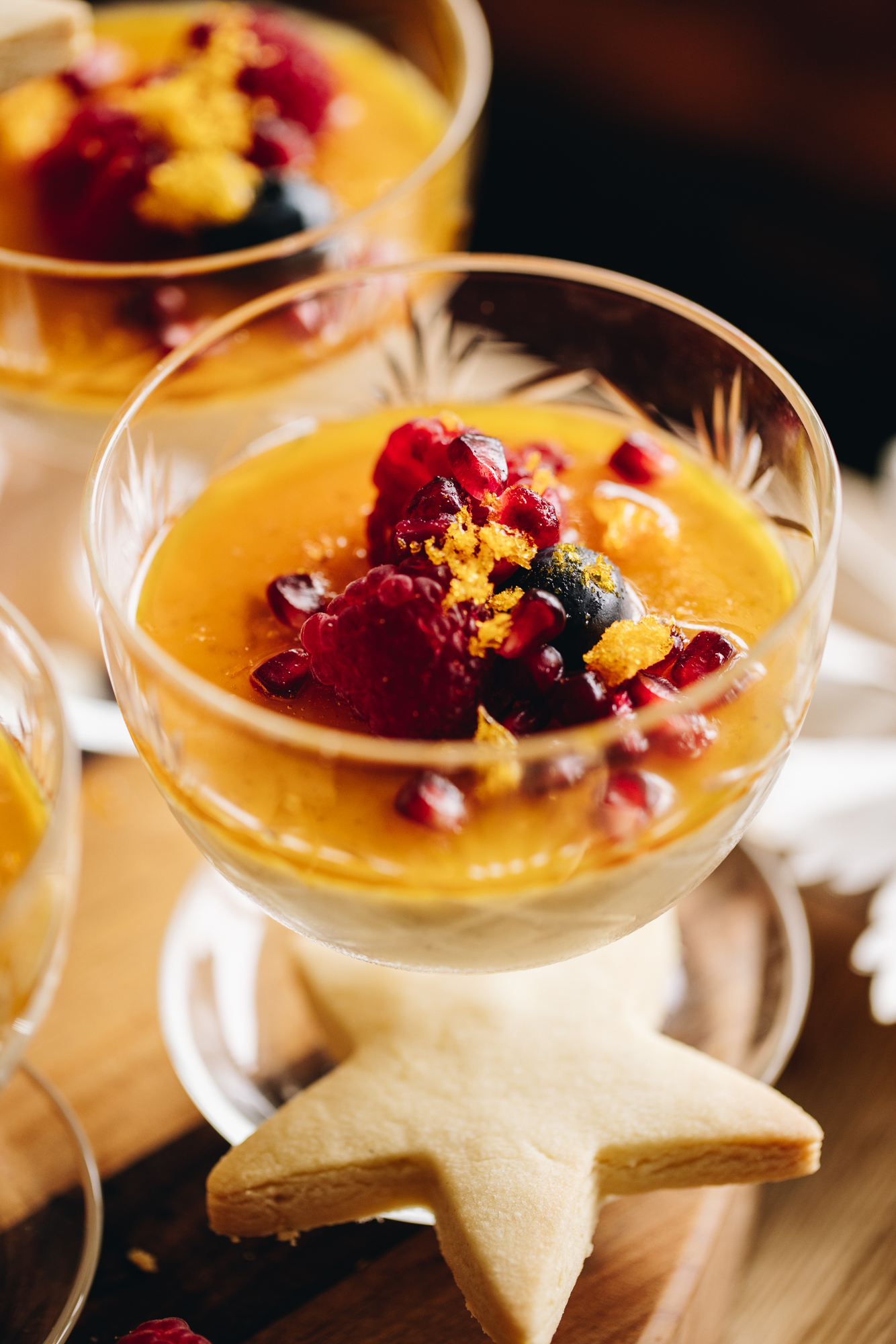 A close up reveals a glass vintage ramekin sitting on a wooden table. It has been filled with orange Panna Cotta with orange syrup, berries and freeze-dried mandarin crumbs on top for decoration. A star shortbread rests on the rim of the glass. 