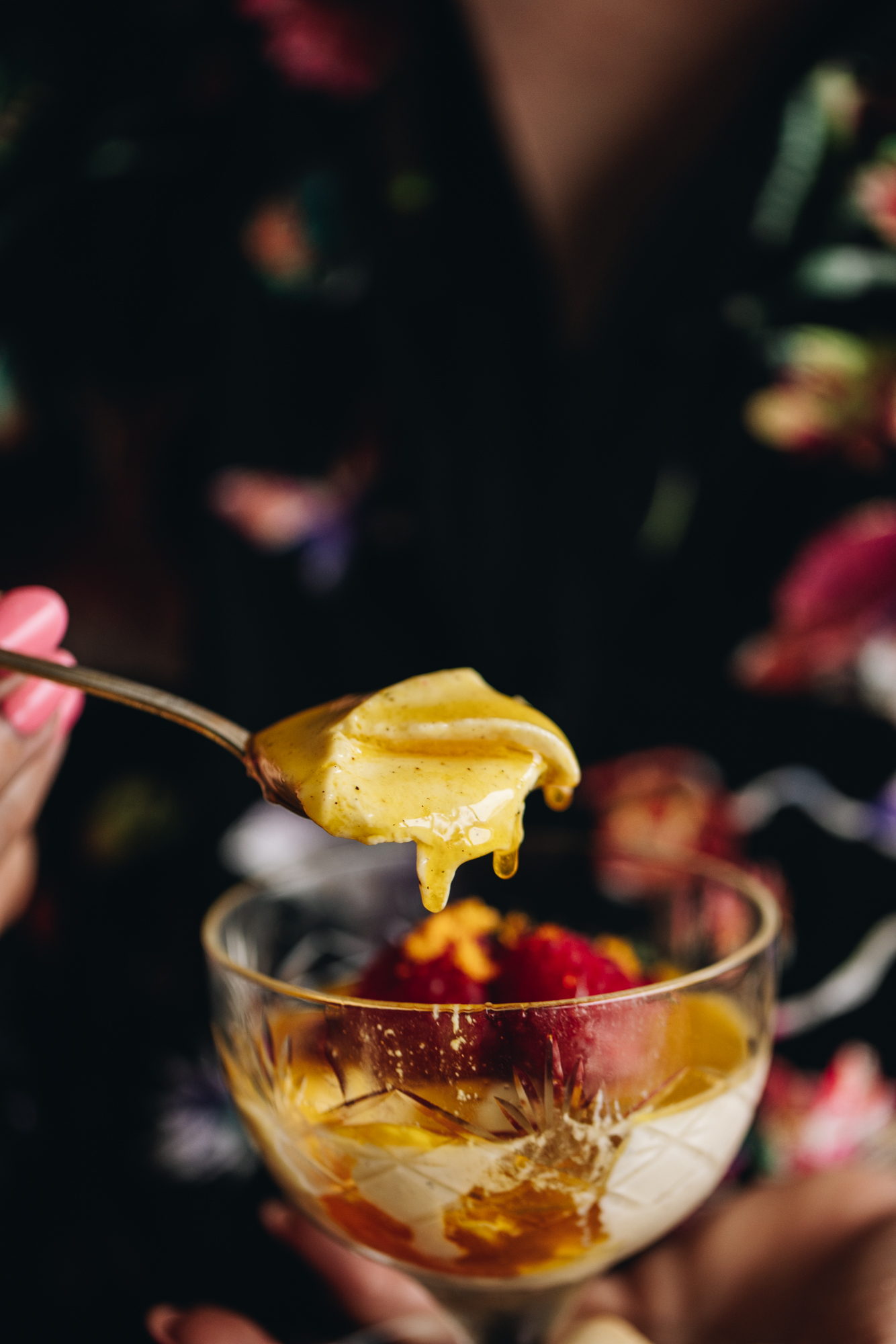 A single Orange Panna Cotta is being held in Naomi's hands. It is a vintage glass with the creamy panna cotta and orange syrup on top of it with fresh raspberries and blueberries. A teaspoon has lifted out a spoonful of the Panna Cotta revealing the soft texture and sticky orange syrup. 