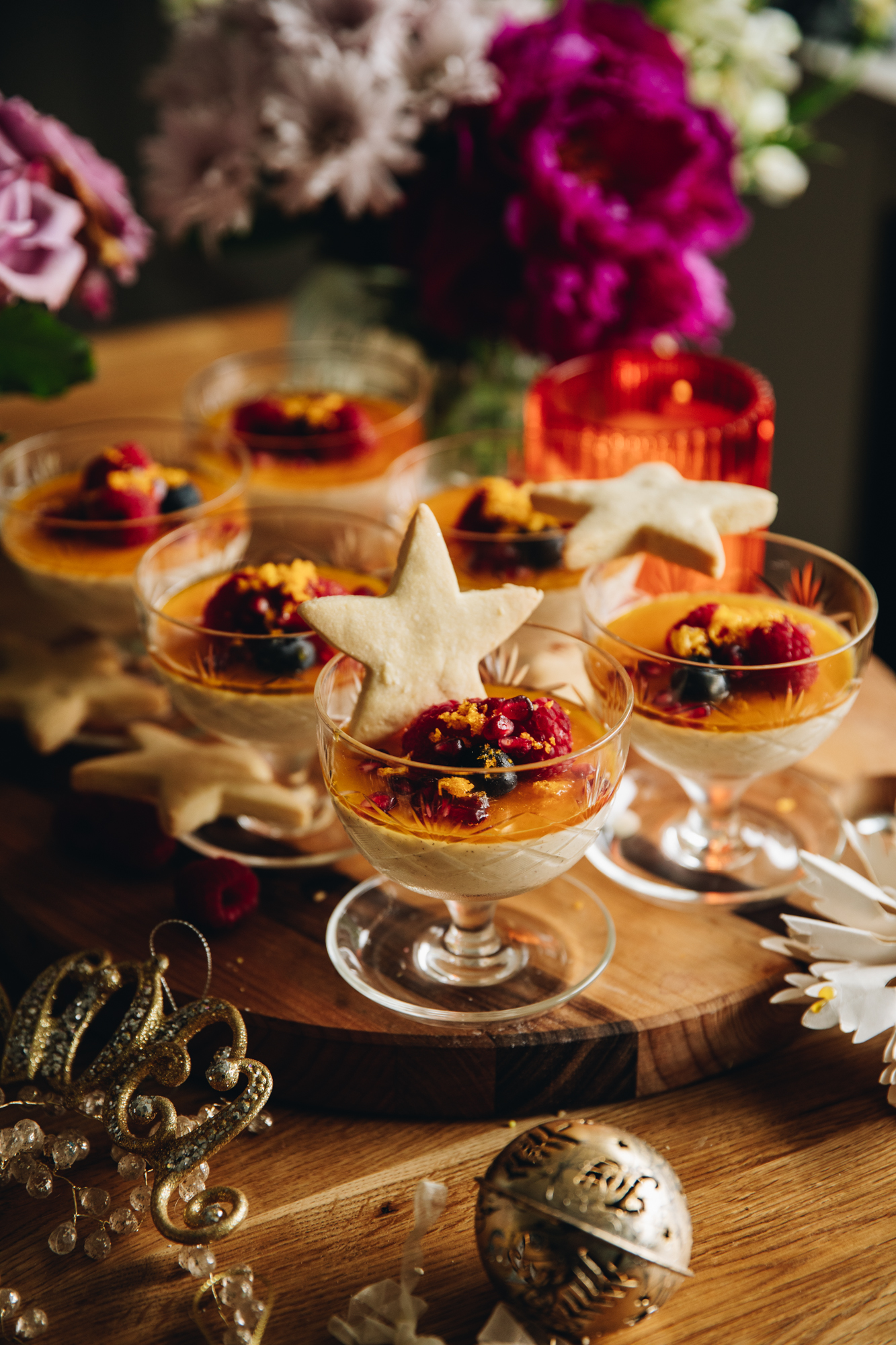 6 glass vintage ramekins sit on a wooden table. All of them have been filled with the orange Panna Cotta with orange syrup, berries and freeze-dried mandarins on top. Shortbread stars are added in to one dessert, another one lays on top of a glass and the others sit on the lower rim of the glass. An orange candle burns in the background and purple flowers are seen.