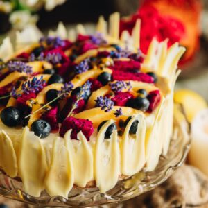 A lemon cheesecake with berries on top is decorated with White Chocolate decorations that are rustic leaf shapes.