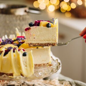 A piece of Lemon and White Chocolate Cheesecake is being lifted from the whole cheesecake. It is suspended in the air with a cake server. It has fresh berries on top with white chocolate decorations.