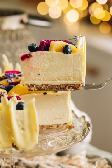 A piece of Lemon and White Chocolate Cheesecake is being lifted from the whole cheesecake. It is suspended in the air with a cake server. It has fresh berries on top with white chocolate decorations.