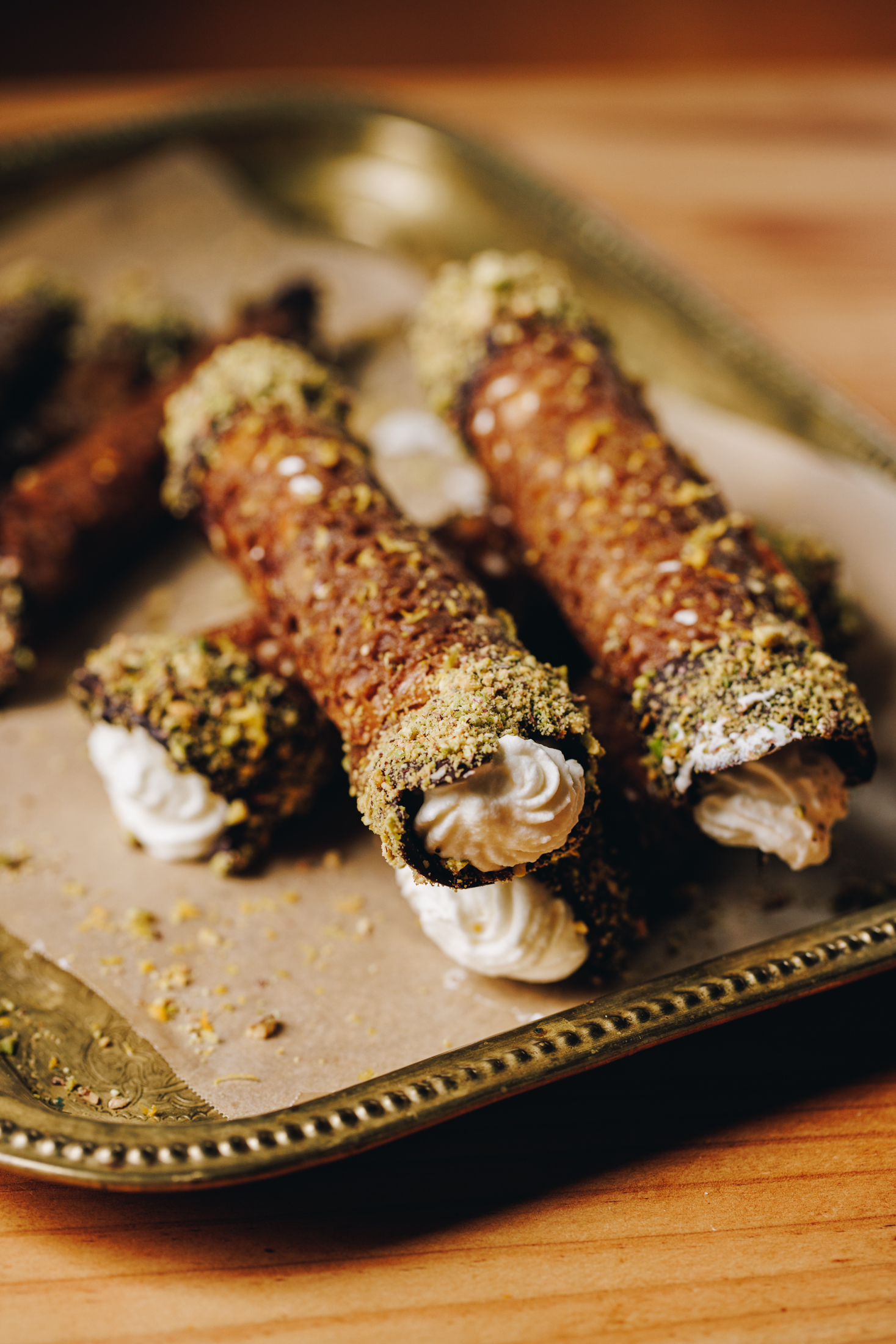 A gold tray lined with brown baking paper sits on a wooden table. On top of the tray is a pile of filled and decorated homemade brandy snaps. The ends have been dipped in dark chocolate and pistachio crumbs and they have been filled with piped cream. 