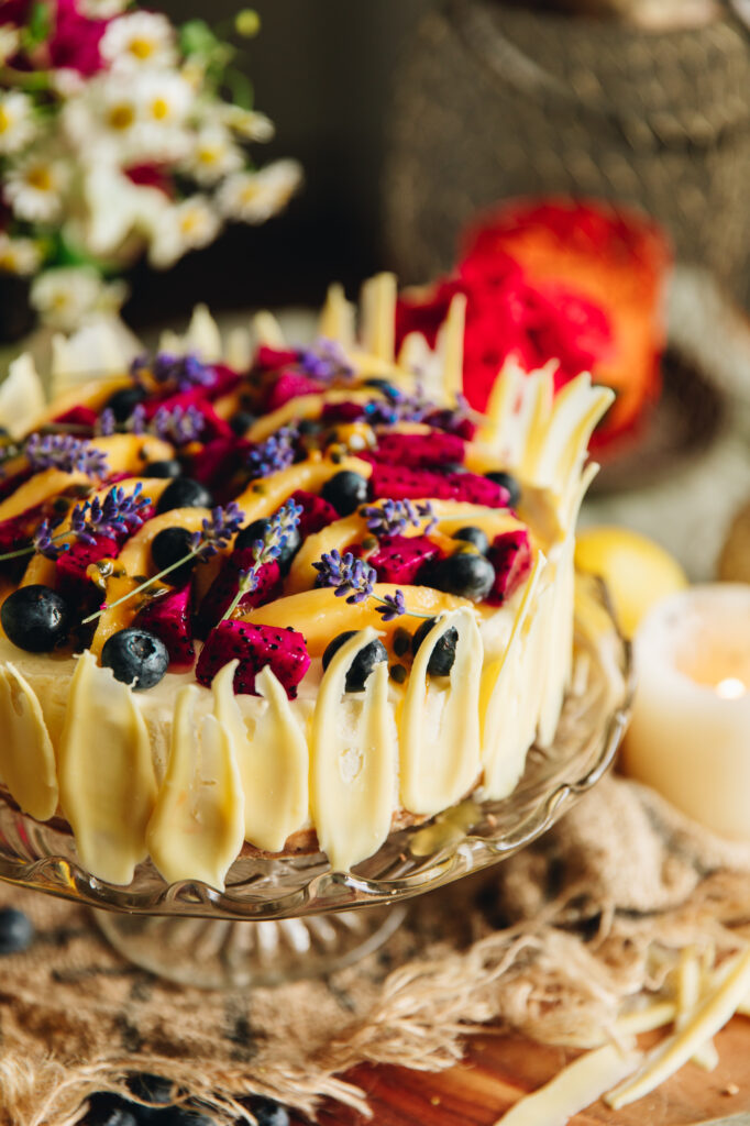 A lemon and white chocolate cheesecake is decorated with fresh fruit on top and fresh lavender flowers. Around the edge of the cheesecake is white chocolate decorations. It is on a glass cake stand.